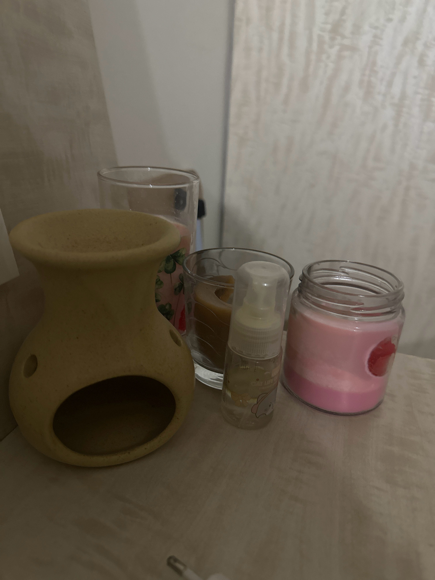 The image displays a collection of decorative items on a light-colored surface. In the foreground, to the left, is a light yellow ceramic oil burner with a hollowed-out base and a rounded top opening. To the right of the oil burner are three containers. The first is a clear glass containing a pink candle with a red strawberry design. Behind this is another clear glass containing a brown candle. Next to the pink candle is a small, clear spray bottle with a white cap, containing a light yellow liquid and a small cartoon elephant sticker on the side. The background consists of a wall with a textured, light-colored finish. This scene is not identifiable as being in Lalitpur, Nepal, or any specific city.