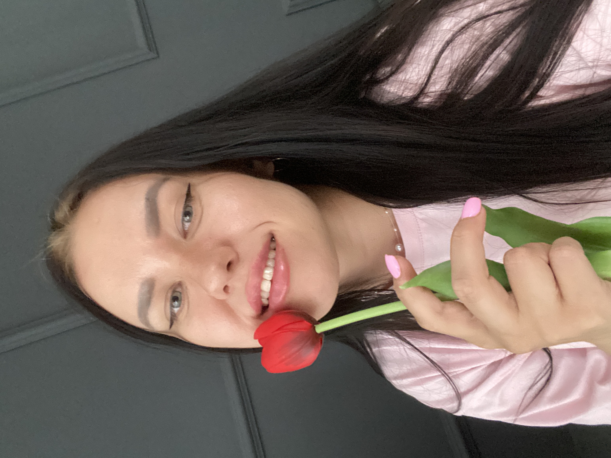 A young woman with long, dark hair and light blue eyes holds a red tulip to her lips. She is wearing a pink t-shirt and a pearl necklace. Her fingernails are painted pink. The background is a dark gray wall with molding. The image appears to be a selfie taken indoors. There is no information available to suggest the location is in Chaikovsky, Russia.