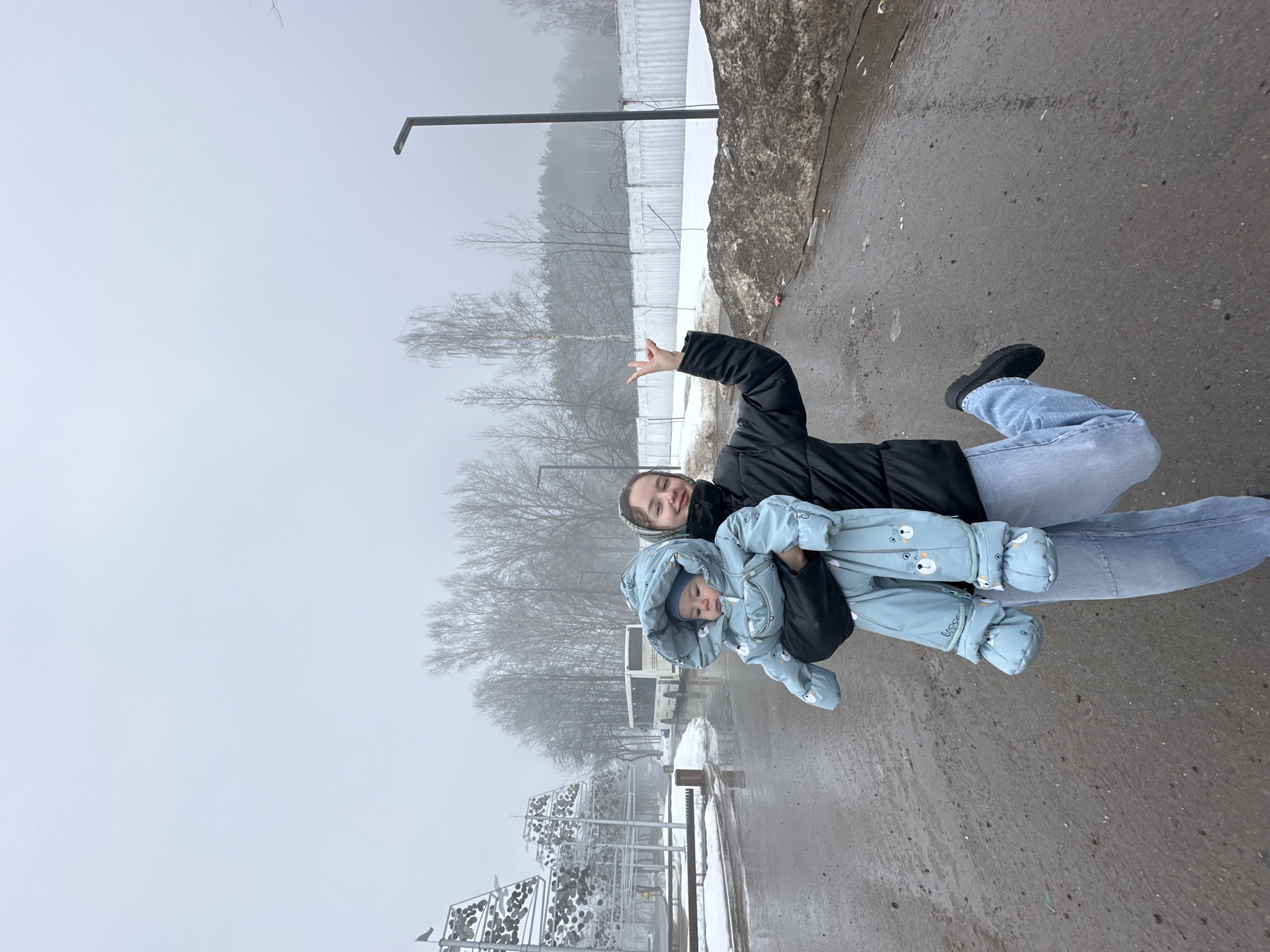 A woman in a black jacket and blue jeans is holding a child in a light blue snowsuit. The woman is smiling and has her hand raised in a waving gesture. The child is looking down. They are standing on a paved surface in an outdoor setting with snow and ice visible. A metal fence, bare trees, and a light pole can be seen in the background. In the distance, there is a white bus and snowy structures. The location appears to be in a park area in Chaikovsky, Russia, on a foggy or overcast day.
