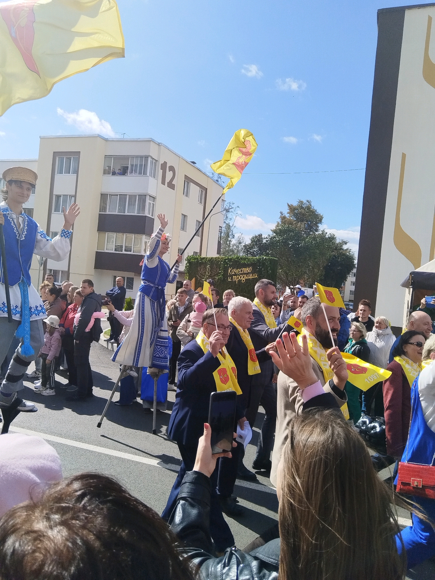A street parade is underway in Brest, Belarus, under a clear blue sky with scattered clouds. Two individuals on stilts, dressed in traditional Belarusian attire, are prominently featured. The individual on the left wears a straw hat and a blue and white embroidered tunic, waving with their right hand. The individual on the right, in a similar blue and white outfit with a floral headdress, is also waving. They are surrounded by a crowd of onlookers of various ages, many holding small yellow flags with a red symbol and the number "1097". Some people are holding phones up, appearing to take pictures or videos. In the background, a modern multi-story building with the number "12" visible on its facade stands adjacent to a building adorned with a large gold abstract design. A hedge with a sign reading "Качество и традиции" (Quality and Traditions) is also visible. The ground is paved with asphalt, marked with a white line. The overall atmosphere suggests a festive event or celebration.