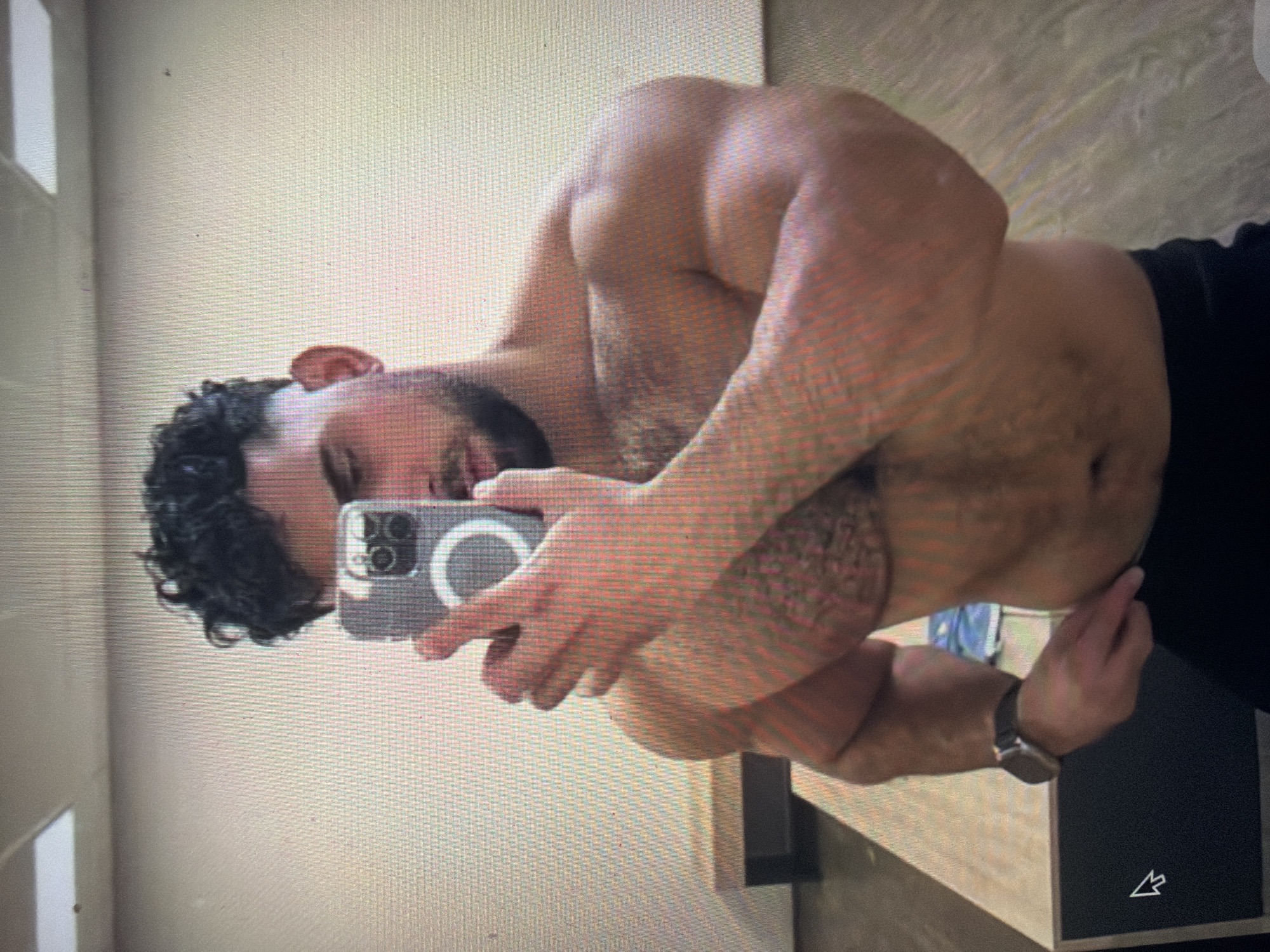 A shirtless male subject, exhibiting a muscular physique with dark, curly hair and a beard, is depicted taking a mirror selfie in an indoor setting located in Shumen, Bulgaria. The subject holds a smartphone with a clear case and a visible MagSafe ring in his right hand, partially obscuring his face. His left arm is bent, with his hand resting near his hip. A watch with a dark strap is visible on his left wrist. He is wearing dark-colored bottoms. The background reflects light-colored walls, rectangular ceiling light fixtures, and a dark floor. A wooden bench or counter with a dark base is visible in the mid-ground. The image itself appears to be a photograph of a screen, displaying a pixel grid pattern and a mouse cursor in the lower-left corner.