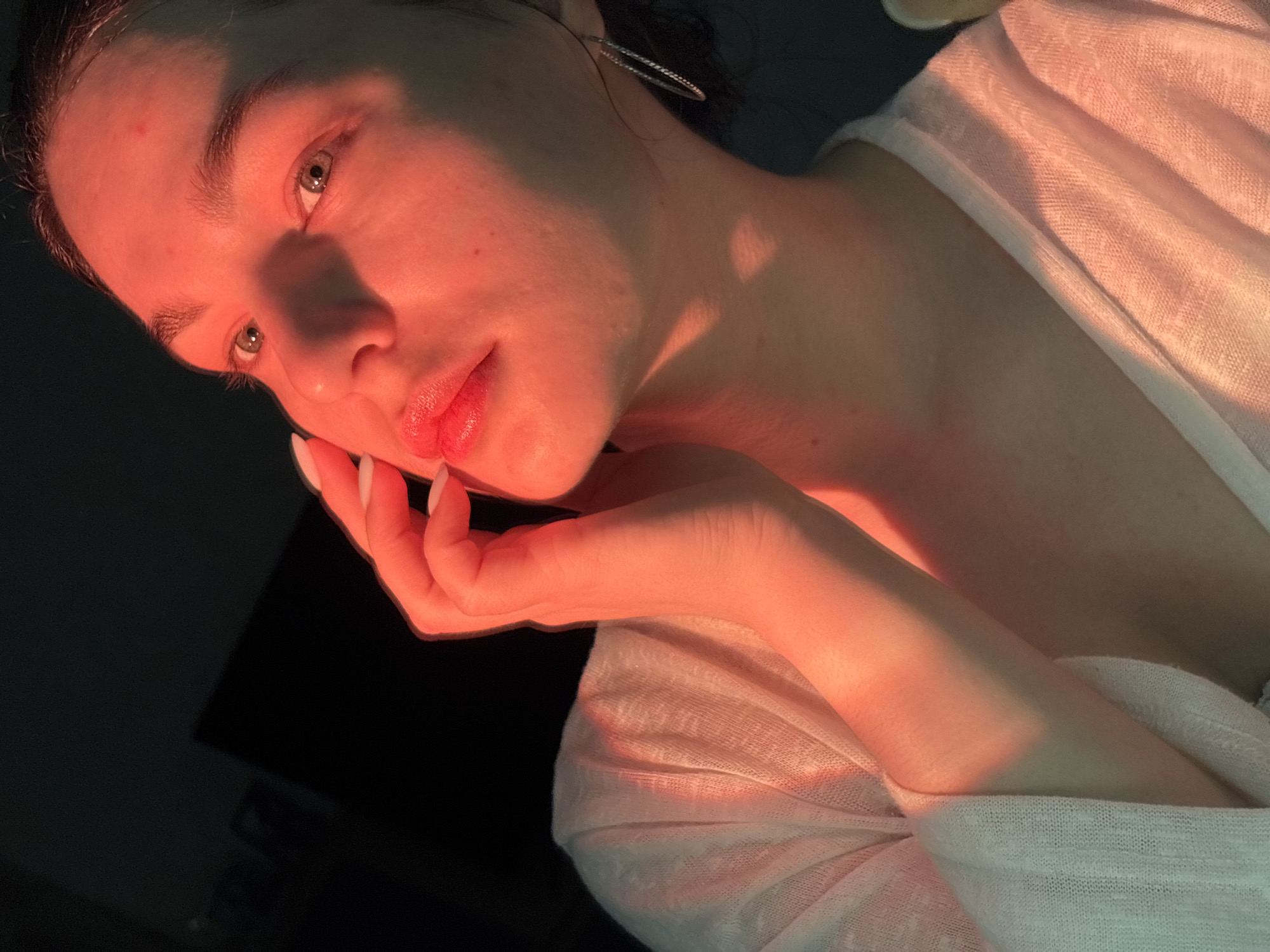 A close-up image shows a light-skinned person with their face and hand illuminated by an orange-red light. The person has light eyes, dark eyebrows, and pink lips, and is wearing a sheer white top. The person's hand is near their face. The background is dark and out of focus, but appears to be a dark surface.
The location in the city of Chaikovsky, Russia is not discernible from the image.