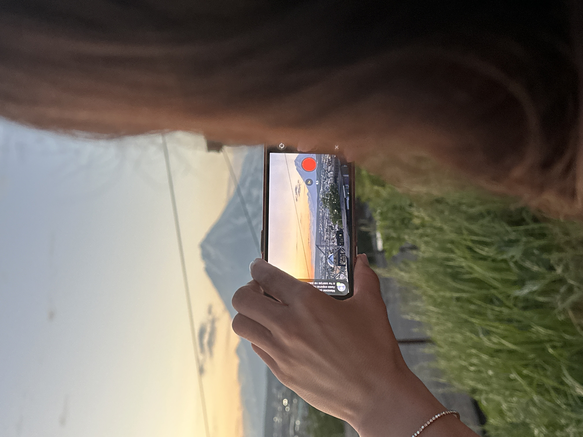 An over-the-shoulder shot shows a person filming Mount Fuji at sunset with their phone. The phone screen displays the mountain, a city, and a red record button. The person's hand, adorned with a bracelet, holds the phone. In the foreground, tall green grass grows. The background features a hazy sky with clouds and the iconic snow-capped volcano. The scene appears to be set in a city like Petropavlovsk-Kamchatsky, Russia, with Mount Fuji captured as if viewed from a distance.