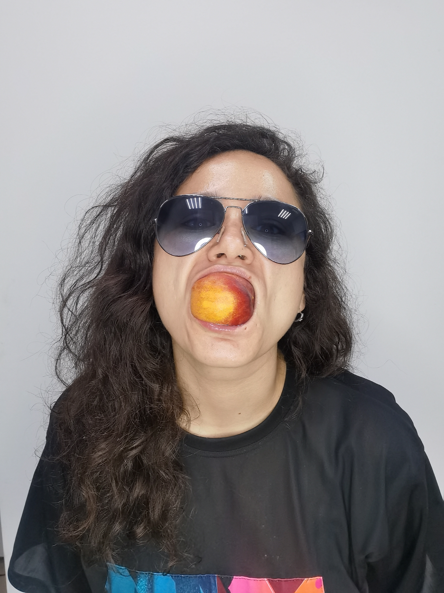 A person with long, dark, curly hair and aviator sunglasses has a peach in their mouth. They are wearing a black t-shirt. The background is a plain white wall. There is no indication of the city of Chaikovsky, Russia in this image.