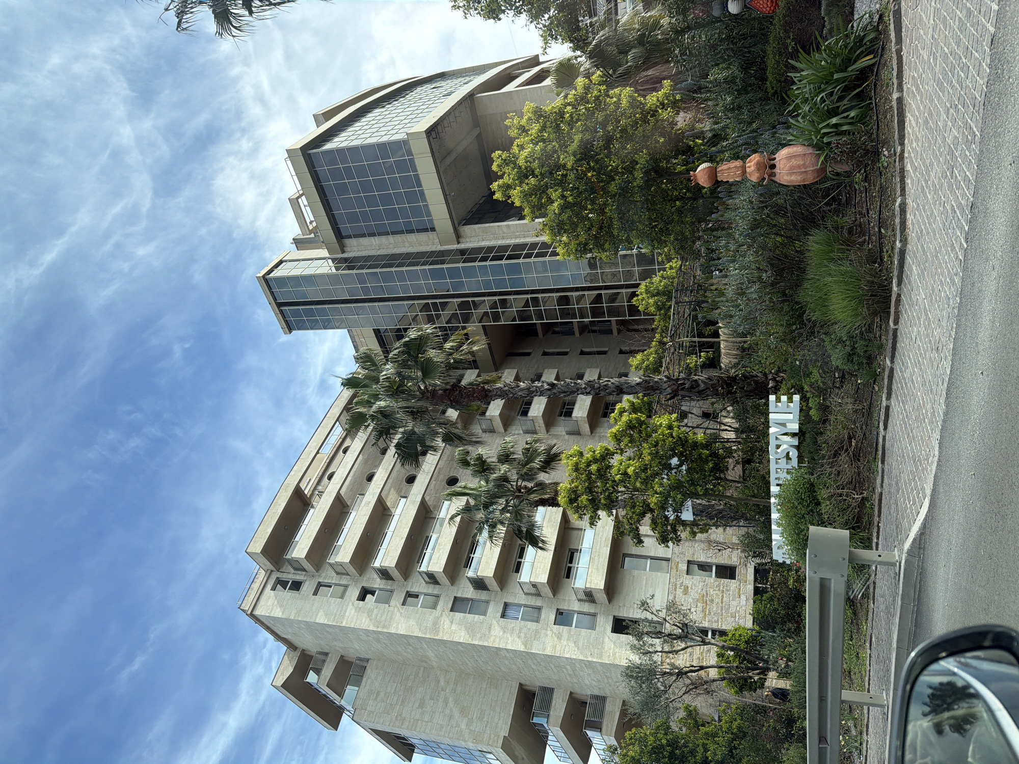 The image shows a low-angle view of a tall building in Budva, Montenegro. The building has a mix of architectural styles, with a lower section featuring stone or light-colored cladding and recessed balconies, and an upper section with large glass windows. Palm trees grow in front of the building. A sign that says "LIFESTYLE" is visible in front of the building. The sky is partly cloudy with a blue hue. A portion of a car's side mirror is visible on the right of the frame.