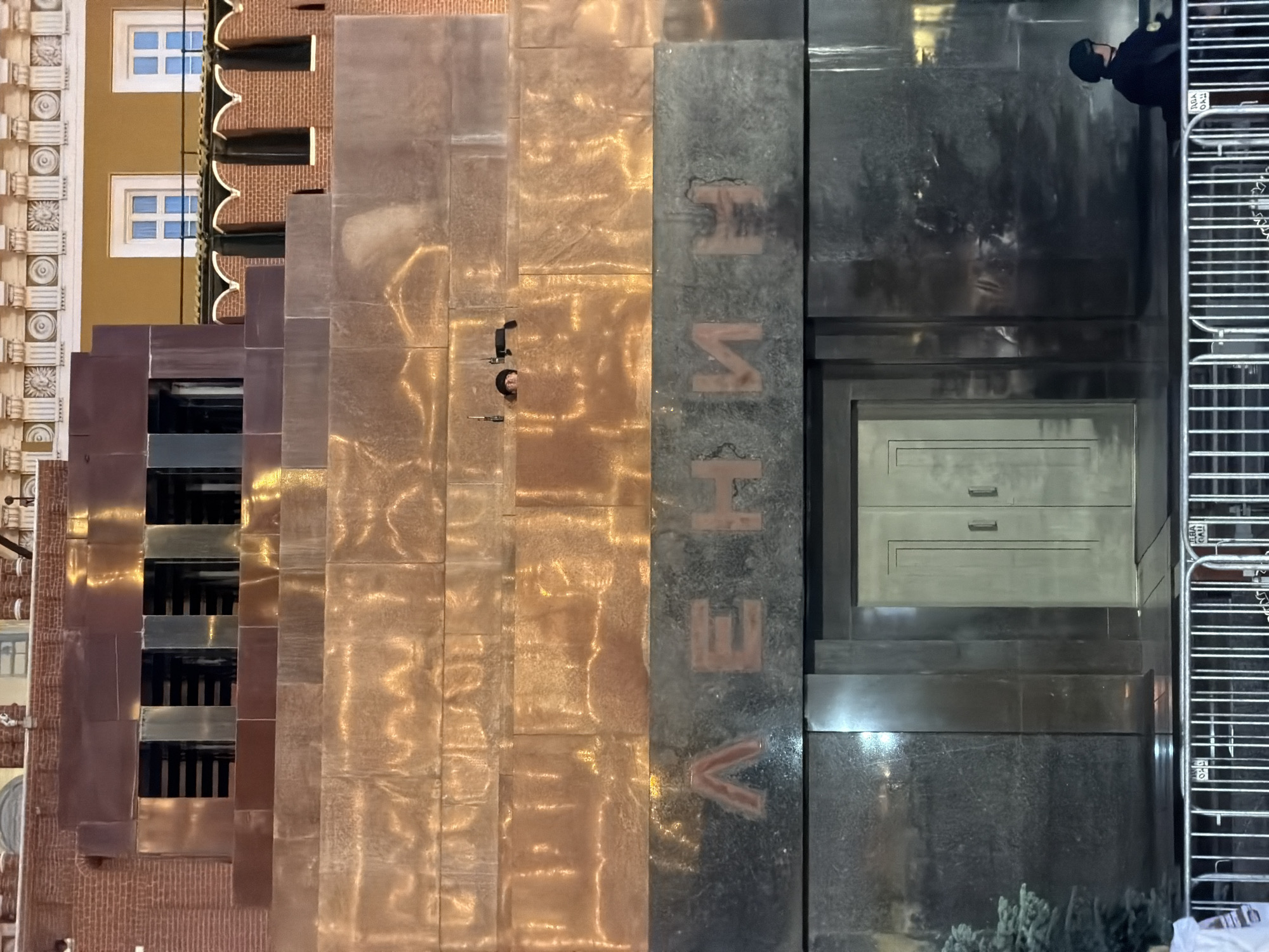 This detailed image captures Lenin's Mausoleum located in Red Square, Moscow, Russia. The mausoleum is a monumental, multi-tiered structure composed of dark, polished stone, with several prominent sections clad in a reflective, bronze-toned material, particularly on its upper and middle levels.
The upper portion of the mausoleum features a dark, stepped section with four rectangular, recessed openings revealing darker interiors. Above and behind this, elements of the Moscow Kremlin are visible, including red brick walls with distinctive crenellated tops and part of an ornate yellow building with white-framed windows and a decorative classical cornice.
Below the bronze-toned cladding, a dark stone band displays the name "ЛЕНИН" (LENIN) in large, stylized red-orange Cyrillic letters. Directly beneath this inscription is the mausoleum's entrance, consisting of a light-colored or metallic double door with vertical recessed panels, set within a dark stone facade.
Two individuals are visible in the scene, suggesting a security presence:
1. On one of the bronze-clad tiers, roughly two levels above the ground and to the right of the mausoleum's central axis, a person's head and shoulders are visible, peeking over the edge. This individual wears a dark hat or helmet, and two communication devices (walkie-talkies) are placed on the ledge beside them.
2. On the ground level, to the far right of the entrance, another person in a dark uniform and helmet is seated with their back partially obscured by a metal barrier.
A silver-colored metal crowd control barrier runs horizontally across the entire foreground. A small, dark green coniferous tree is positioned on the ground level to the far left. The reflective surfaces of the mausoleum prominently display ambient light reflections, indicating either night conditions with strong artificial illumination or low natural light.