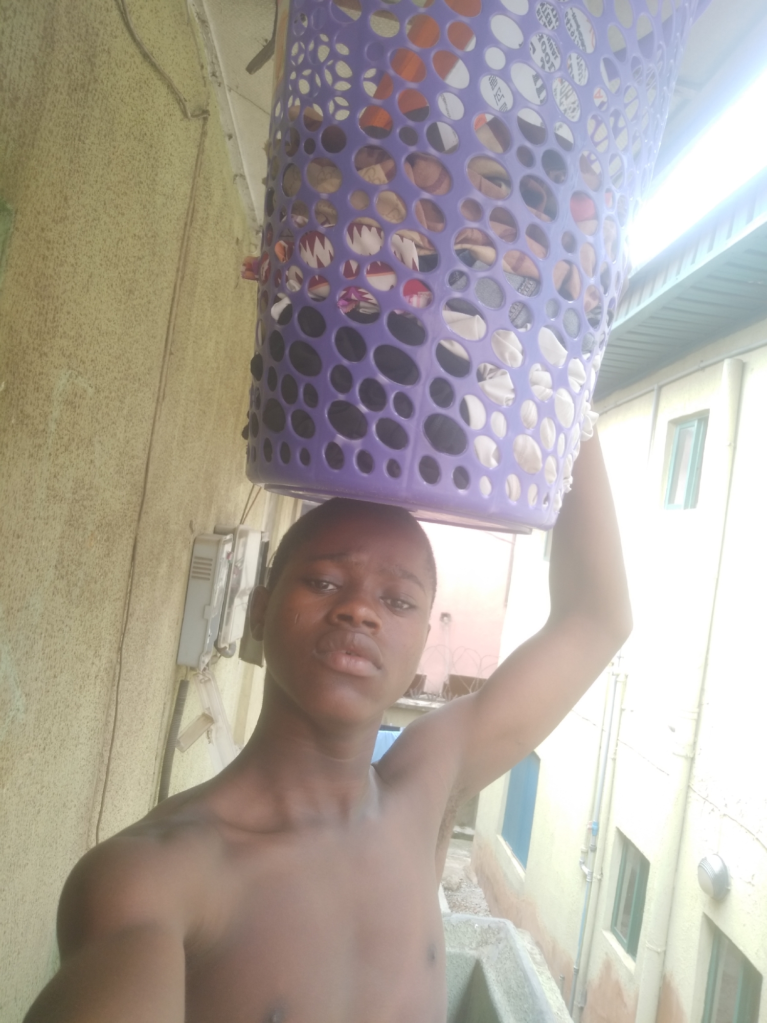 A shirtless young male, possibly of African descent, stands outdoors, holding a purple laundry basket filled with clothing on his head. He appears to be standing in an alleyway or narrow space between buildings. The buildings on either side are painted in light colors. Utility lines and pipes are visible along the walls. An electrical meter is also visible on the wall behind him. The location appears to be in an urban area, possibly Shangisha, Nigeria.