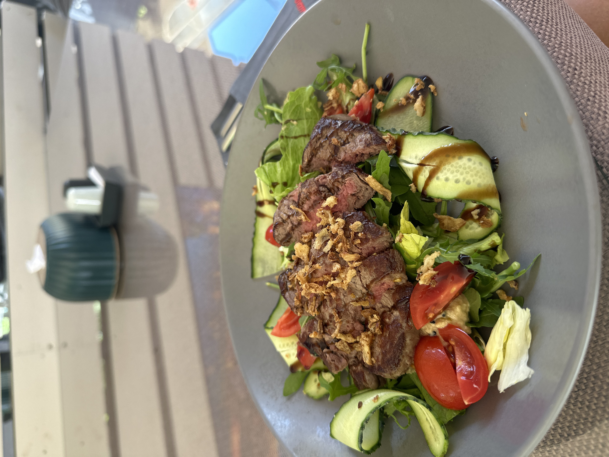 The image shows a close-up of a salad with steak on a gray plate. The salad contains sliced cucumbers, cherry tomatoes, and leafy greens. The steak is sliced and topped with crispy fried onions. A dark sauce, likely balsamic glaze, is drizzled over the salad and steak. The plate is on a table with a woven placemat. In the background, slightly out of focus, is a teal-colored tissue holder and a salt and pepper shaker. The setting appears to be an outdoor restaurant or cafe. The image was taken in Shirokaya Schel, Russia.