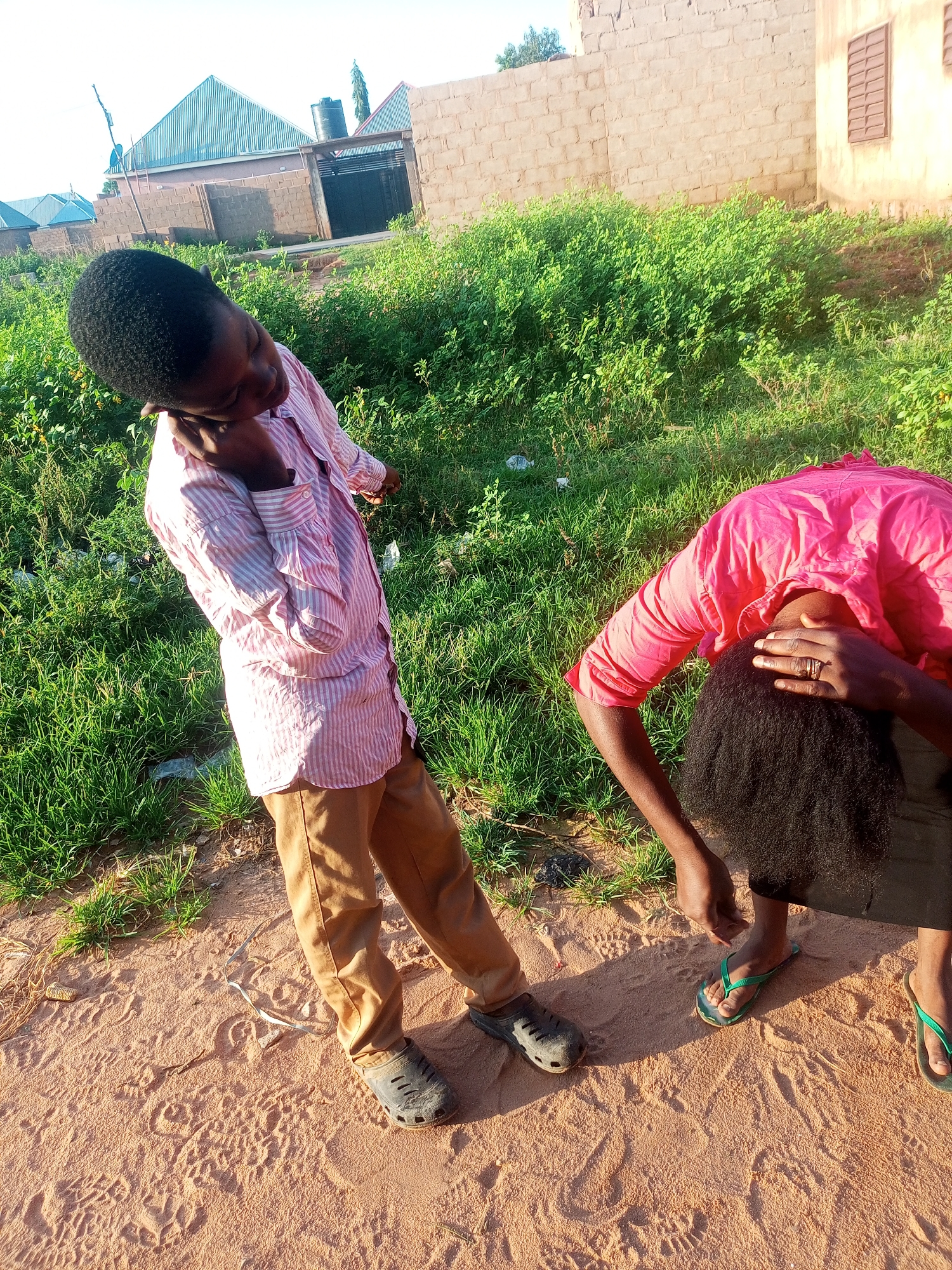 In Tempure, Nigeria, two individuals are present in an outdoor environment characterized by sandy ground and lush green vegetation. The person on the left, a young male, is positioned in a slightly contorted pose, leaning back with his head tilted, his right hand touching his ear, and his left arm bent at the elbow. He is wearing a pink and white striped long-sleeved shirt and tan trousers, with black shoes on his feet.
The second individual, positioned to the right and slightly further back, is bent over with their head obscured by their arms, which are resting on their head. This person is wearing a bright pink long-sleeved shirt and dark trousers, with green flip-flops on their feet. A ring is visible on the right hand of this individual. The background shows a brick wall, structures with corrugated metal roofs, and dense green foliage. The scene appears to be illuminated by natural daylight.