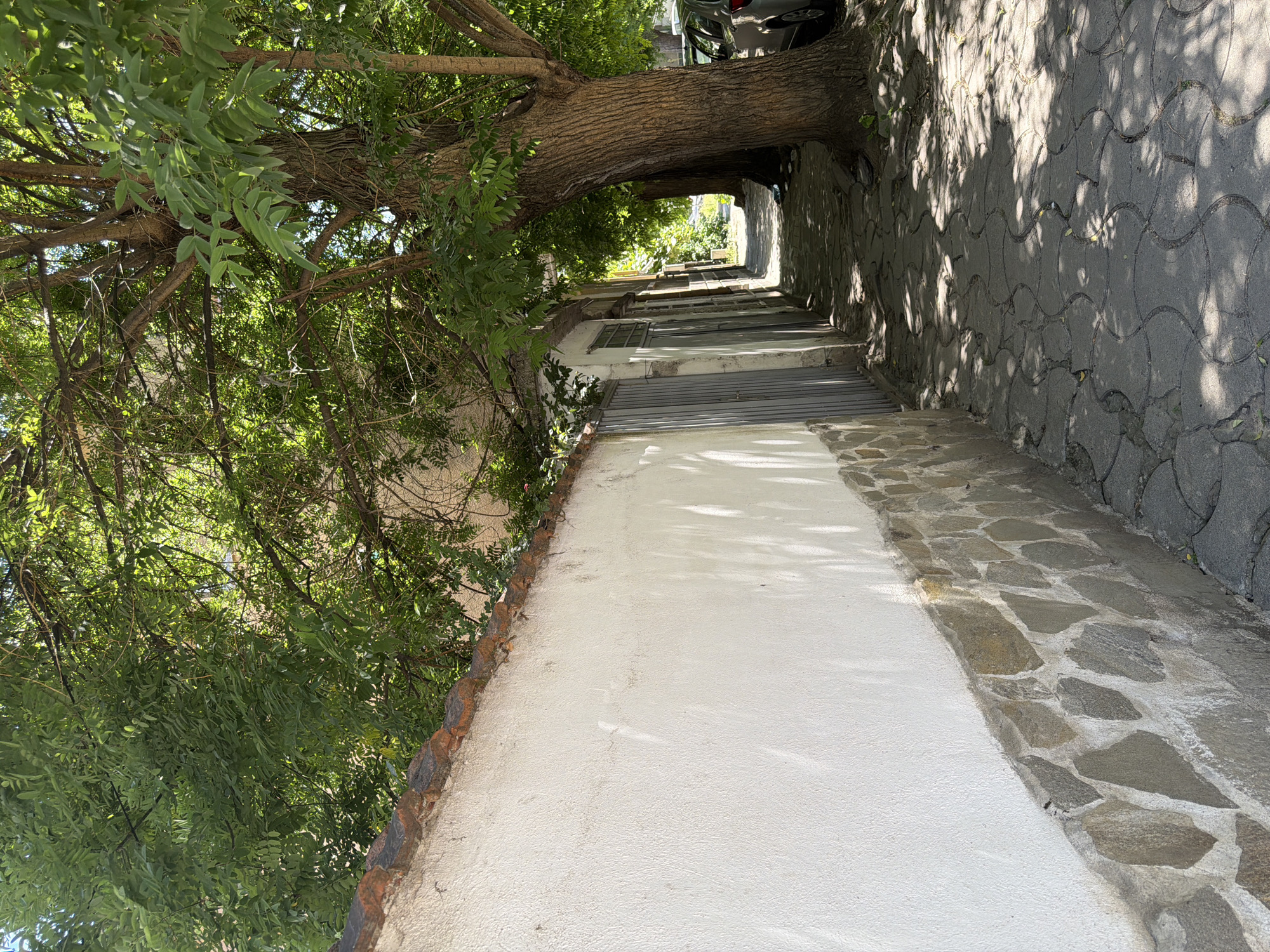 The image depicts a narrow, tree-lined sidewalk in Pazardzhik, Bulgaria, adjacent to a stone-and-plaster wall. Sunlight filters through the dense foliage of a large tree on the right, casting dappled shadows on the patterned cobblestone path. To the left, the wall is made of rough, light-colored plaster with a base of irregular stones. Beyond the wall, several gray metal doors are visible, suggesting a residential or commercial area. Further down the path, more trees and the corner of a parked silver car can be seen. The scene appears peaceful and ordinary, characteristic of a sunny day in a European town.