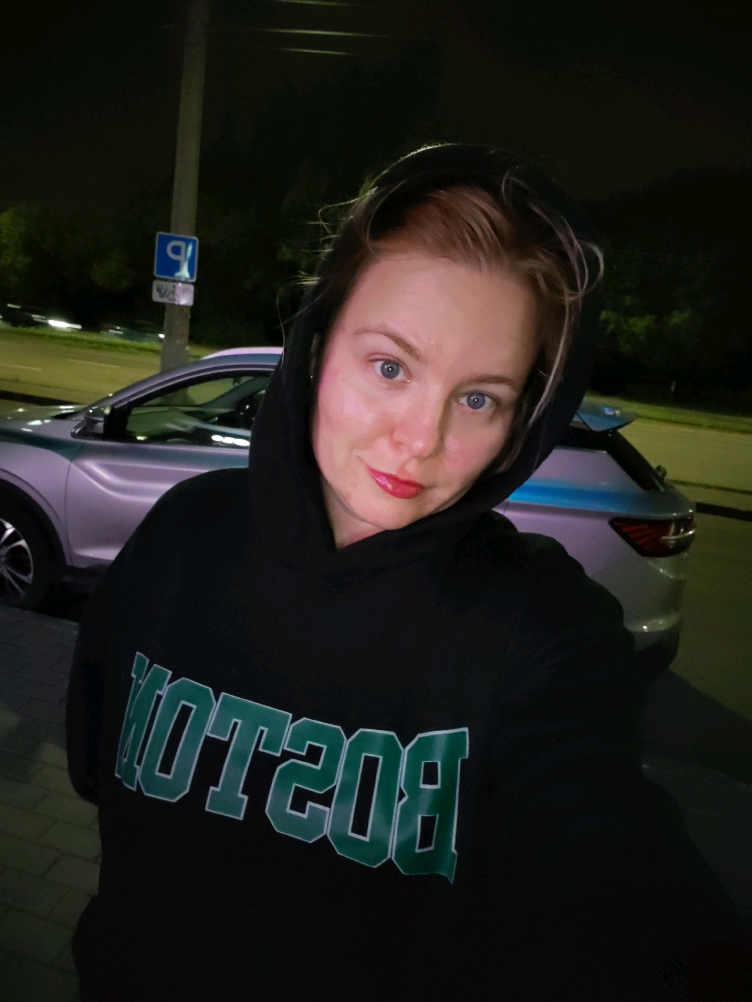 A woman with blonde hair and blue eyes, wearing a black hoodie with "BOSTON" printed on it in green and white letters, is taking a selfie at night. She is standing outdoors, with a silver car parked behind her. In the background, there is a street light and trees. A blue sign with a white symbol is visible on a pole. The scene appears to be in Izhevsk, Russia, likely on a street.