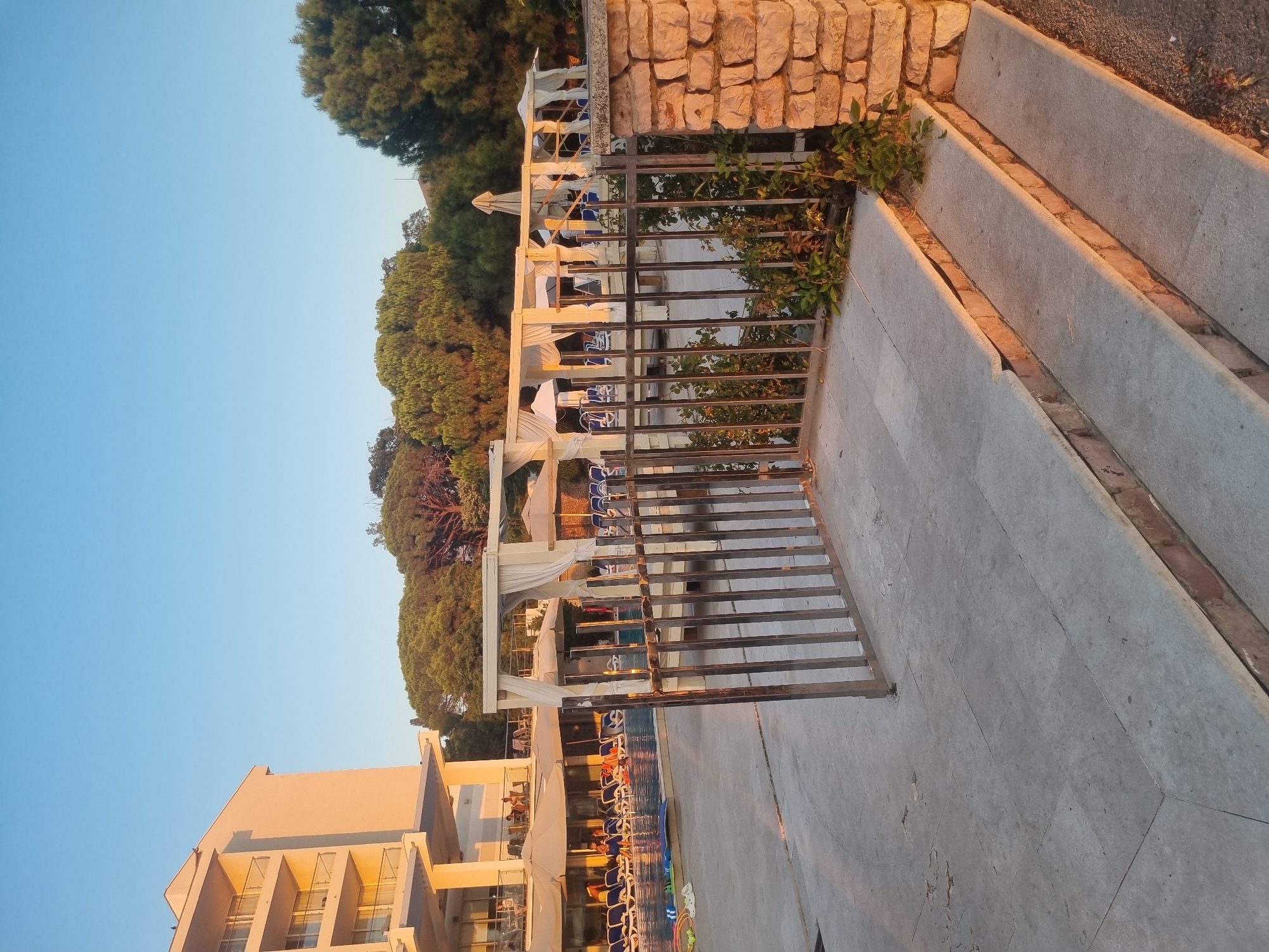 This image depicts a resort area at sunset. A white hotel building with multiple balconies is visible on the left. In the center, a swimming pool is surrounded by lounge chairs and umbrellas. Several white canopies with draped fabric are set up near the pool. A metal gate with vertical bars is in the foreground, partially obscuring the view of the pool area. To the right of the gate is a stone pillar. Green trees form a backdrop behind the resort structures. The sky is clear and blue, indicating good weather. The scene appears to be located in Bar, Montenegro, given the context provided.
