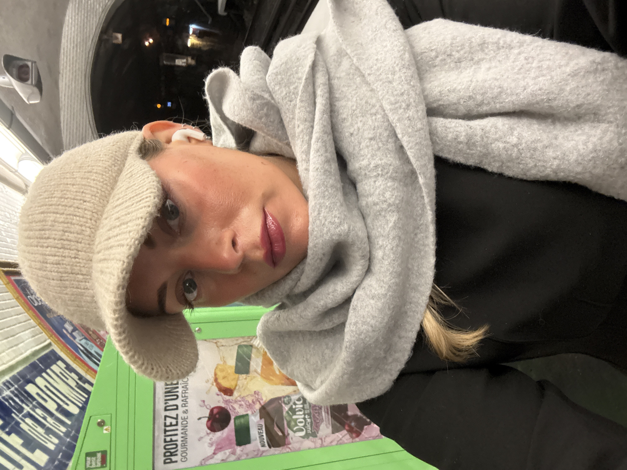 A young woman wearing a light beige knitted cap, a large light gray scarf, and a black jacket is taking a selfie. An AirPod is visible in her right ear. She is standing next to a green vending machine in what appears to be a subway station. The vending machine has a poster with the words "PROFITEZ D'UNE" followed by "GOURMANDE & RAFRAÎCH" and a picture of a Volvic Juicy drink. The background suggests she is on a train or in a subway station. The location, inferred from the signage and vending machine contents, is most likely Paris, France.