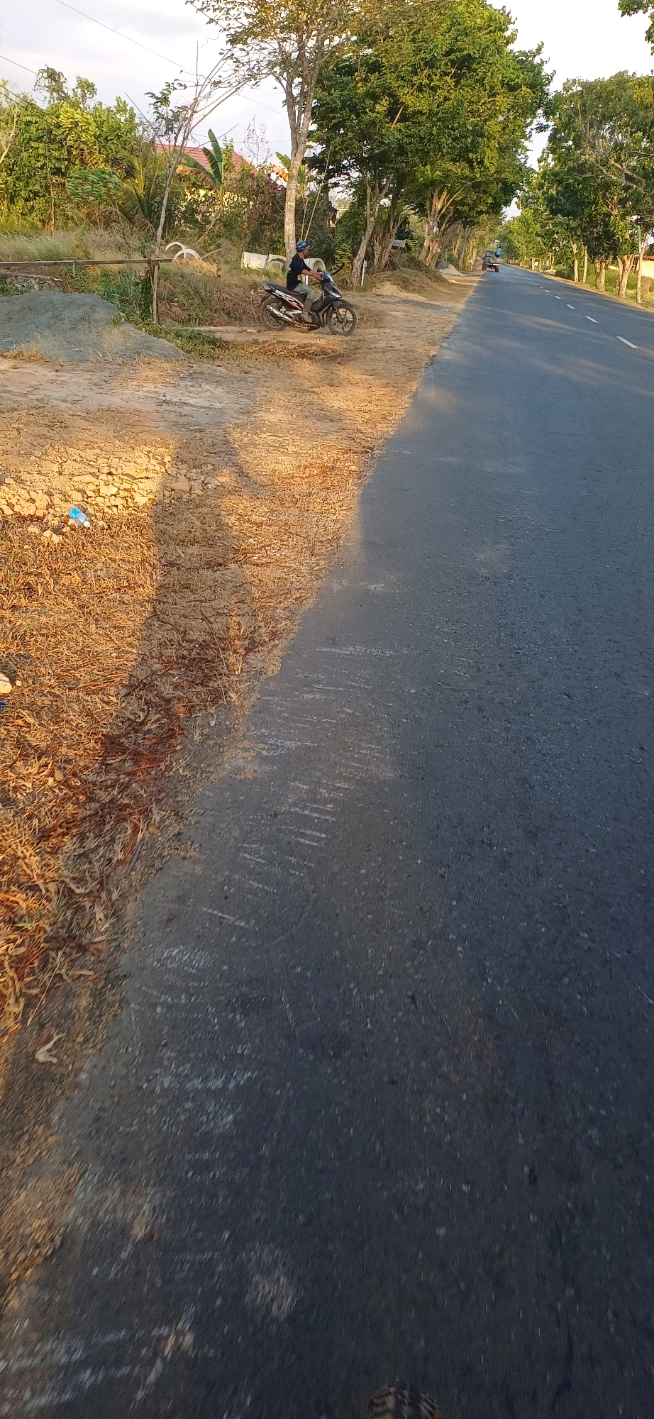 The image depicts a roadside scene in Kersik Putih, Indonesia, during daylight hours. A person wearing a dark shirt and a blue helmet is seated on a dark-colored motorcycle, positioned on the sandy shoulder of an asphalt road. The motorcycle appears to be stationary. The roadside is characterized by dry grass and some scattered debris. Trees with lush green foliage line the road, and a building with a red roof is visible in the background, partially obscured by vegetation. The road itself is asphalt and appears to be a two-lane road with a white line marking the center. The overall environment suggests a rural or semi-rural setting. The sunlight casts shadows on the ground, indicating the time of day. In the distance, other vehicles are visible on the road, suggesting traffic.