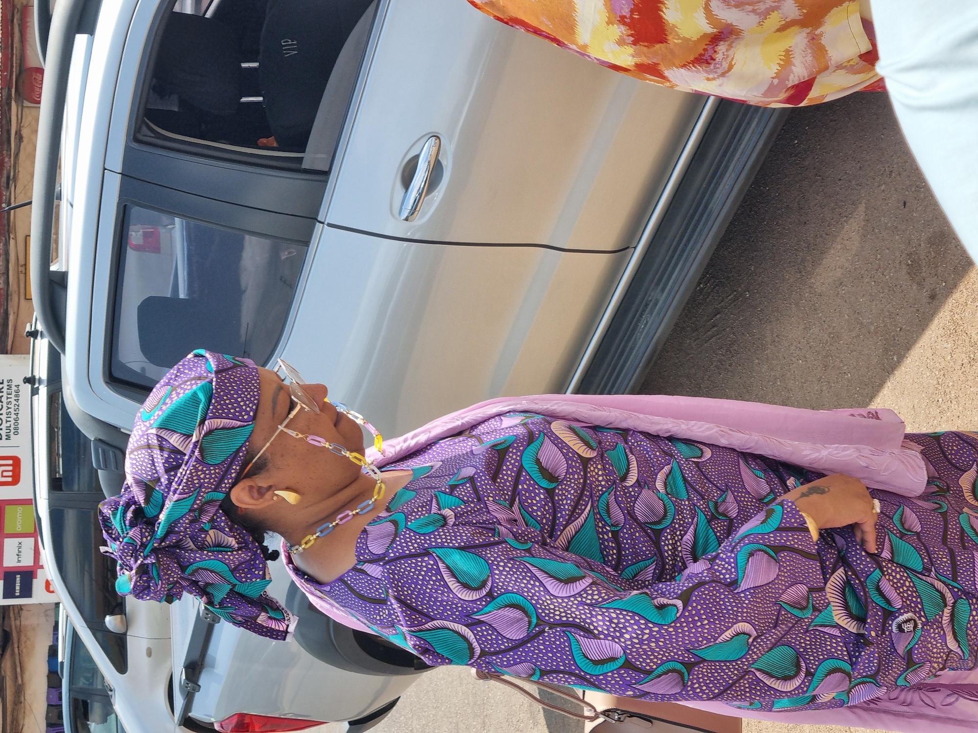 In Jos, Nigeria, a woman wearing a purple and teal patterned dress and headwrap stands next to a silver SUV. She has a pink scarf draped over her shoulders and is wearing a gold necklace and earrings, as well as glasses with a chain. The SUV has a "VIP" sign visible in the window. A sign for "MULTISYSTEMS" with brand logos is visible in the background.