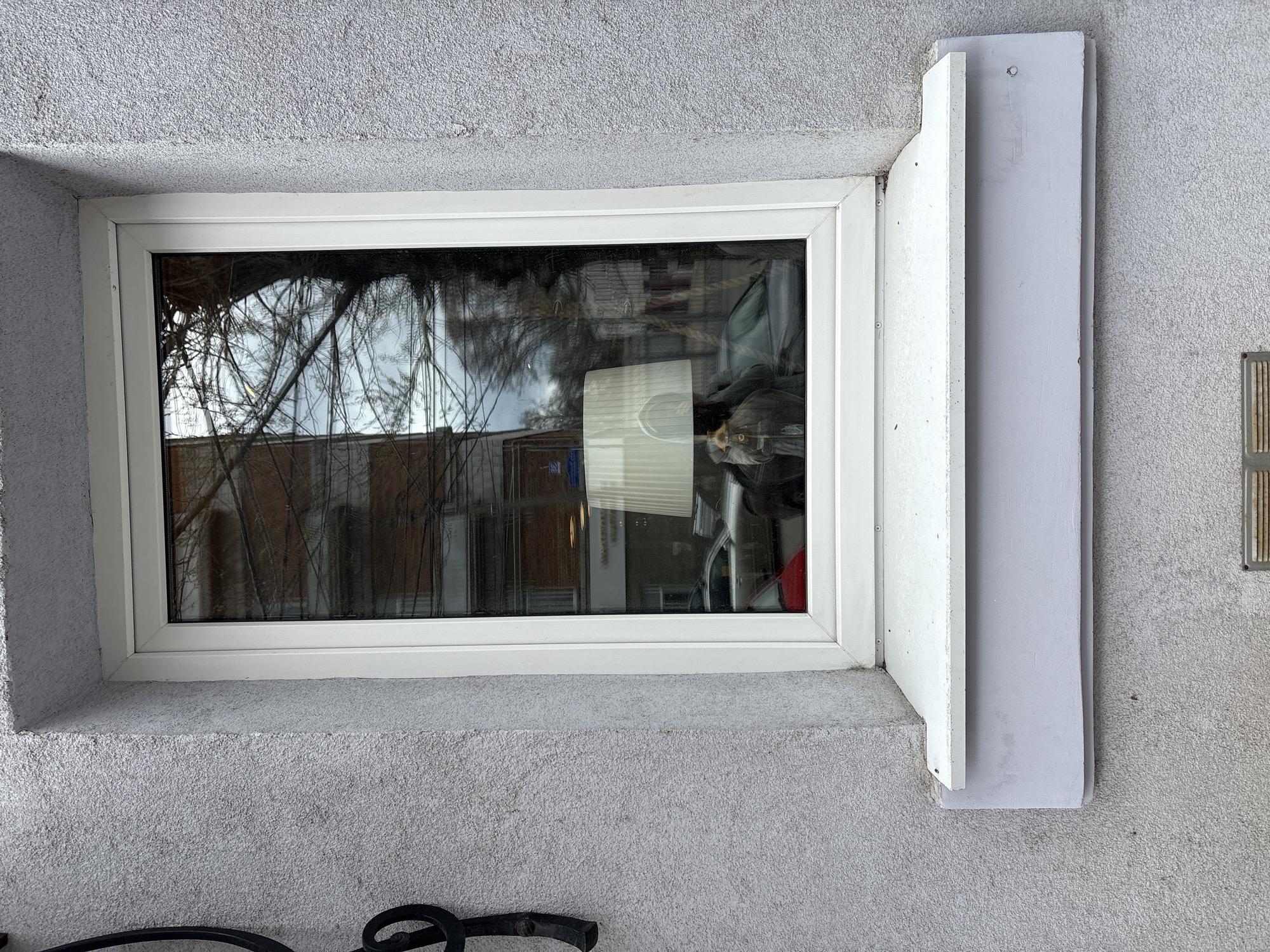 A ground-level view shows a white-framed rectangular window set in a gray stucco wall in Ulyanovsk, Russia. The window reflects a blurred outdoor scene, including trees and a building across the street. To the right of the window, there is a white, vertical structure attached to the wall. Below the window, part of a black metal object is visible.
