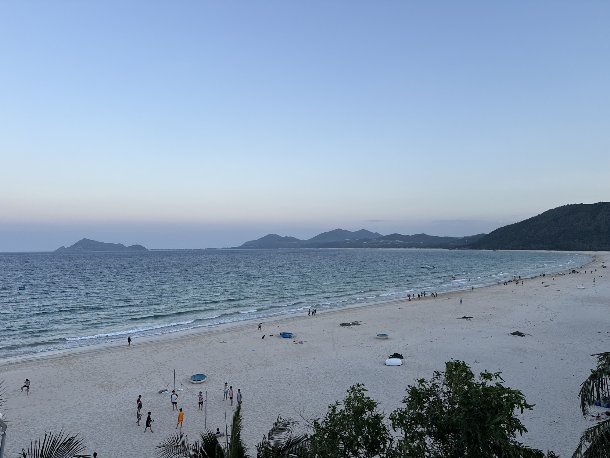 The image captures a wide, sandy beach in Song Cau, Vietnam, during what appears to be the late afternoon or early evening. The sky is a pale, clear blue, transitioning to a lighter hue near the horizon. A serene, turquoise ocean stretches across the frame, with gentle waves lapping the shore. In the distance, a chain of dark, lush green mountains forms the coastline, with some smaller islands visible further out at sea.
Numerous people are scattered across the beach, engaged in various activities. Some are walking along the water's edge, while others are playing or socializing on the sand. A group of young men are playing a game of volleyball near the foreground, with a net set up. Several small, blue and white boats are visible in the shallow water, and some people are wading or swimming. In the foreground, palm fronds and green foliage frame the scene, suggesting the presence of tropical vegetation. The overall atmosphere is peaceful and relaxed, depicting a typical scene of recreational activity on a Vietnamese beach.