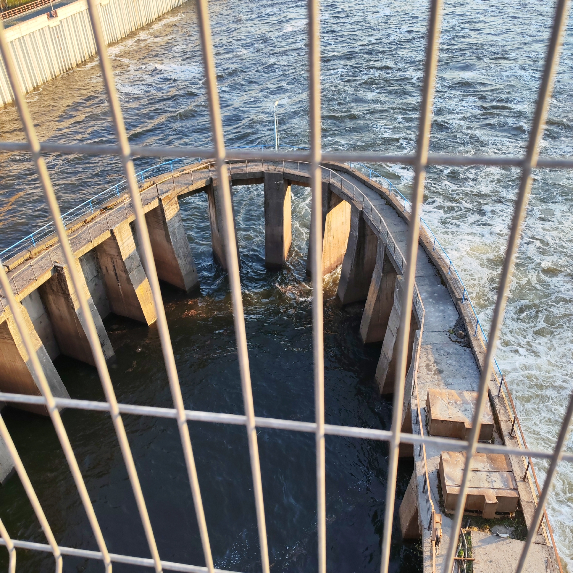 The image shows a curved concrete structure with supporting pillars in a body of flowing water. The structure has a walkway with railings along its perimeter. The water is turbulent and foamy, particularly on the right side of the image. The photograph is taken through a metal fence. In the background, on the left side of the image, there is a concrete wall. According to information available online, this is a view of the Dam in Chaikovsky, Russia.