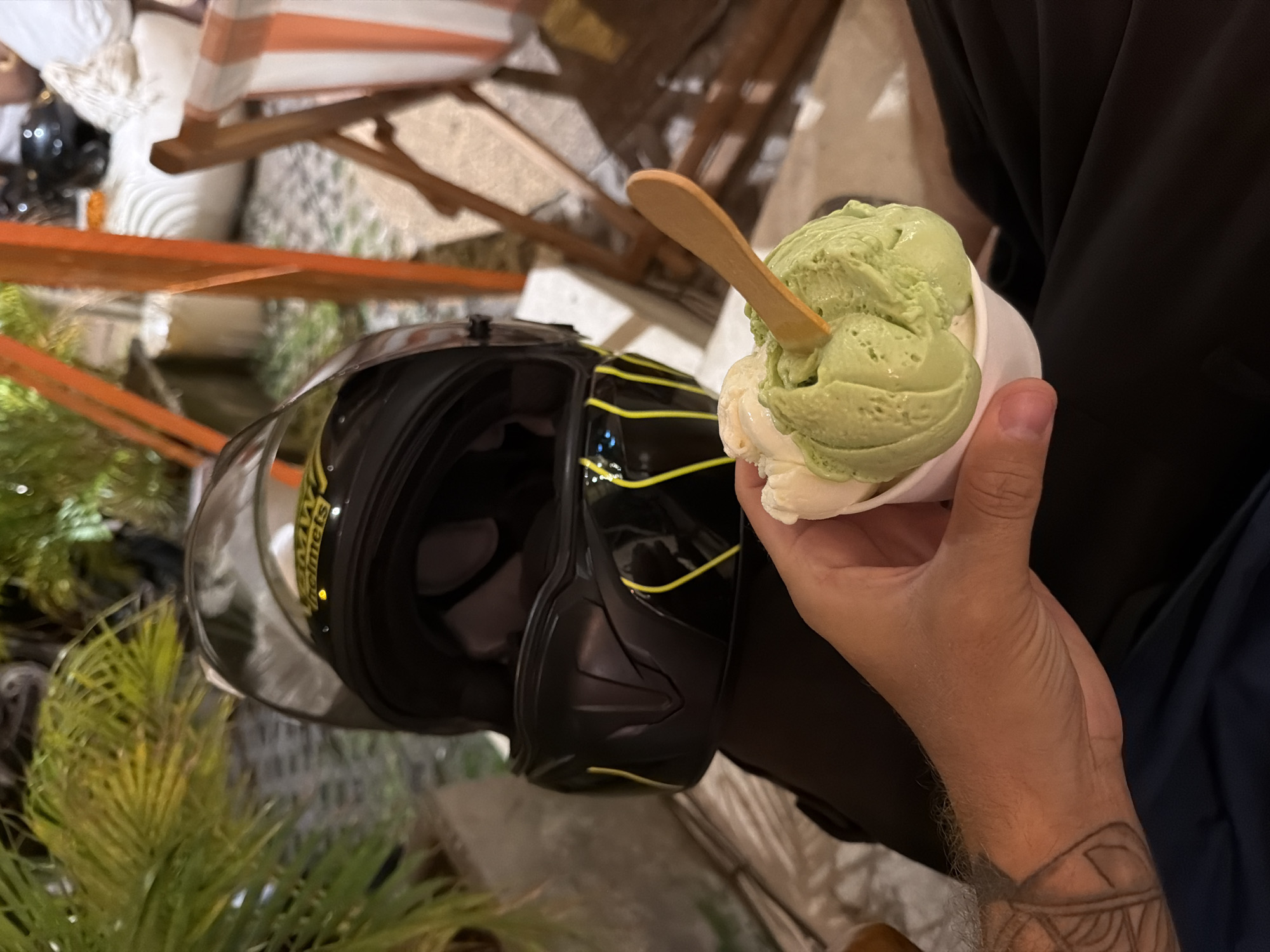 The image, rotated correctly, features a person's right hand holding a white paper cup containing two scoops of ice cream. One scoop is a bright green, possibly pistachio or matcha, and the other is white, likely vanilla. A small wooden spoon is embedded in the green ice cream. The person's forearm shows some hair and a partially visible tattoo.
To the left of the ice cream, a black motorcycle helmet is visible, featuring distinct yellow stripes and a dark visor that is down. The brand name "BMW Helmets" is partially visible on the side of the helmet.
The background is softly blurred, showing green foliage, possibly palm fronds, in the foreground and mid-ground, suggestive of a tropical setting. Behind the helmet and hand, there are blurred wooden structures and hints of light-colored, possibly striped, fabric, indicating an outdoor or semi-outdoor seating area. The lighting suggests it is daytime, perhaps late afternoon. The scene captures a moment of enjoyment and refreshment, likely in North Kuta, Indonesia, with the helmet indicating the person may have recently been on a motorcycle.