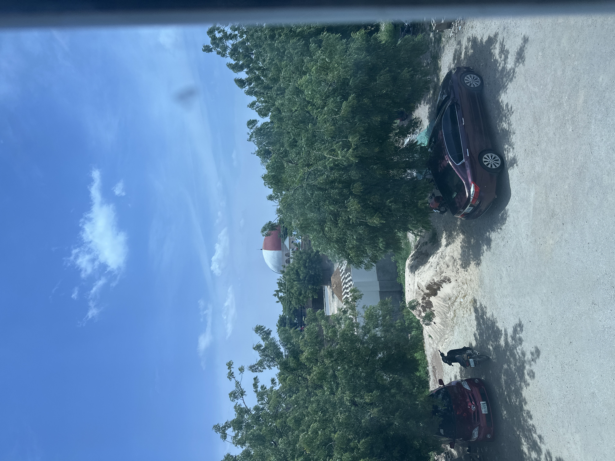 This image captures an outdoor scene with a clear blue sky and scattered white clouds. In the foreground, a dusty, light-colored ground surface is visible, with shadows cast by trees. To the left, a red sedan with license plate ABJ6328 is parked under a tree. Next to it, a dark-colored motorcycle is also parked. In the center of the image, a large mound of sand and debris is present. Further back, a maroon sedan is parked, and to its right, there is a building with a white wall and a dark roof. Beyond that, a distinctive building with a red and white domed roof, resembling a stadium structure, stands against the sky. The overall environment appears to be a somewhat undeveloped urban or semi-urban area, possibly near the Muhammadu Dikko Stadium in Nigeria, given the visual cues of the stadium-like structure and the vehicles.