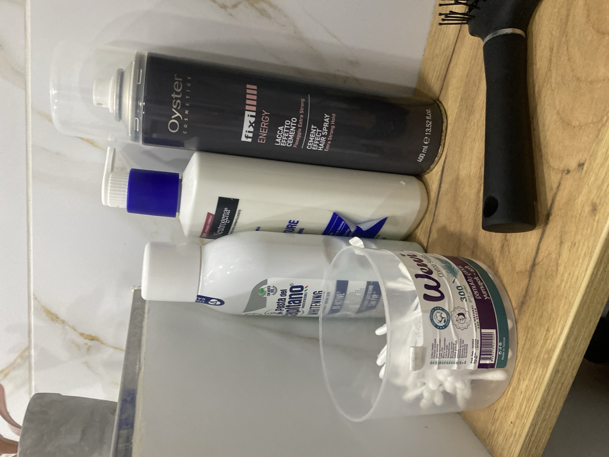 The image shows a shelf in what appears to be a bathroom. On the shelf are several personal care products. From left to right, there is a container of cotton swabs, a bottle labeled "Pasta del Capitano WHITENING", a pump bottle of Neutrogena body lotion, and a can of hairspray labeled "Oyster COSMETICS fix ENERGY". To the right of the hairspray, a black hairbrush with a black handle is partially visible. The background consists of a tiled wall with a marble pattern. The overall lighting suggests an indoor setting. There is no discernible indication of the city of Chișinău, Moldova.