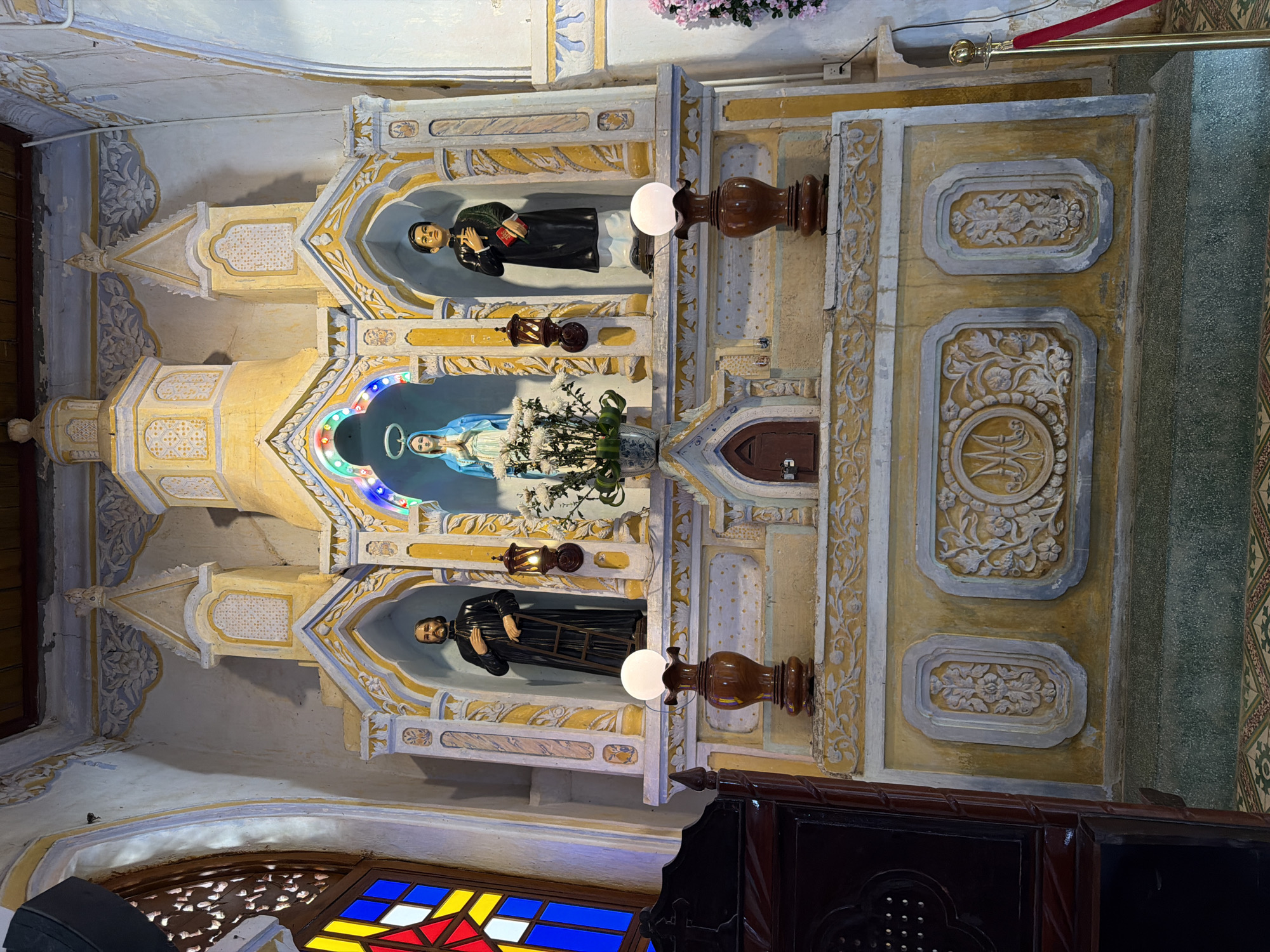 The image depicts an altar within a church in Hội Tín, Vietnam. The altar is a multi-tiered structure, predominantly yellow with white ornamental carvings. At the center of the altar, a statue of the Virgin Mary, wearing a blue robe, is positioned under a canopy adorned with colorful lights. Flanking the Virgin Mary are two statues of male figures in religious attire. To the left, a figure in a black robe stands holding a ladder. To the right, a figure in a black robe holds a red book.
Below the central statues, two wooden lamps with white spherical globes on ornate bases are placed. The lower section of the altar features decorative carvings, including a circular emblem with initials that appear to be "IM". The surrounding walls are painted in muted colors and show signs of age. To the left of the altar, a stained-glass window with red, yellow, and blue panes is visible. In the foreground to the left, a dark wooden furniture piece, possibly a pew or confessional, is partially in view. The floor is covered with patterned tiles. A red velvet rope barrier is present on the right side of the image. The overall scene suggests a religious setting with traditional architectural and decorative elements.