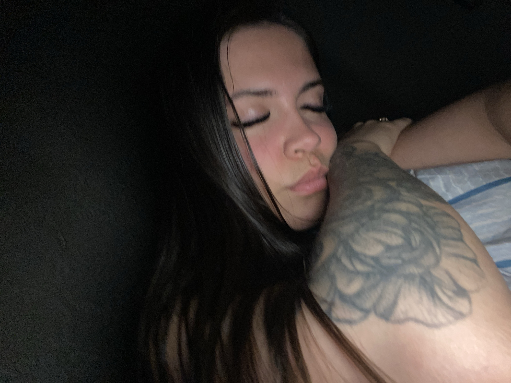 An adult female is depicted sleeping or resting in a dimly lit indoor environment, located in the city of Chaikovsky, Russia. She is positioned on her left side, with her head resting on her folded arms, and her face turned slightly towards the viewer. Her eyes are closed, and her expression is peaceful. She has long, dark hair, which is partially visible around her head. A large, greywash floral tattoo, resembling a rose, is prominently displayed on her left upper arm. A ring is visible on the ring finger of her right hand, which is partially obscured by her head. The surrounding area is dark, with a light-colored, striped fabric, likely a bedsheet, visible beneath her left arm. The illumination is low, casting soft shadows and highlights.