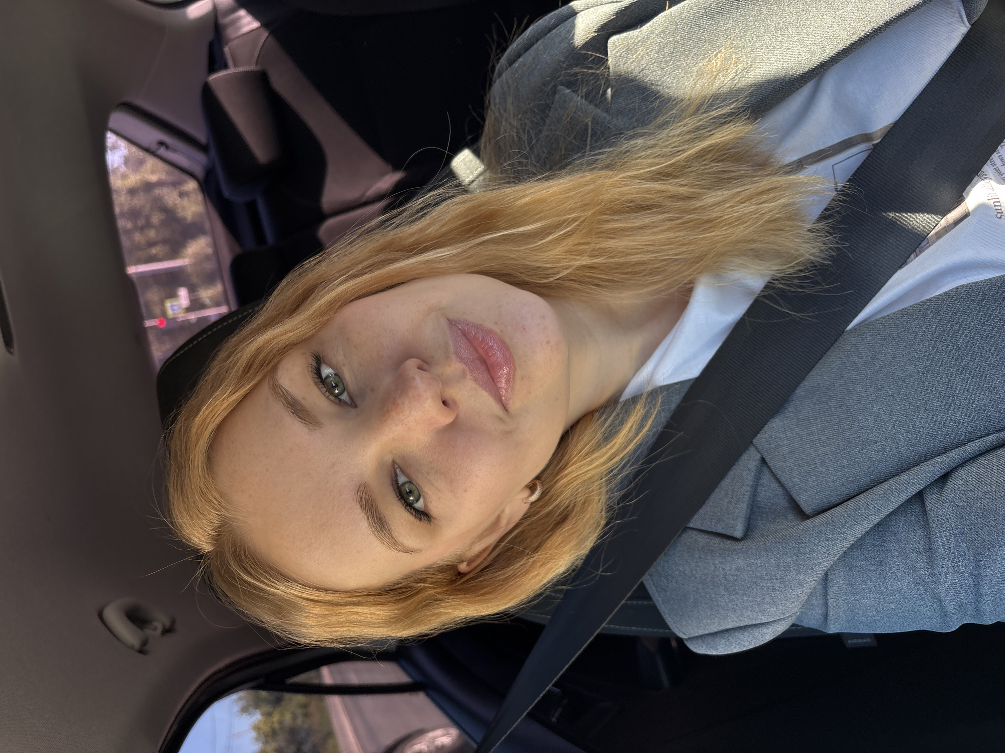 A young woman with blonde hair, green eyes, and freckles is sitting in the passenger seat of a car. She is wearing a grey blazer over a white t-shirt. A black seatbelt is across her chest. The background shows the interior of the car, including the back seats and headrests, as well as a blurred view of trees and a traffic light outside the window. The location is the city of Zhukovo, Russia.