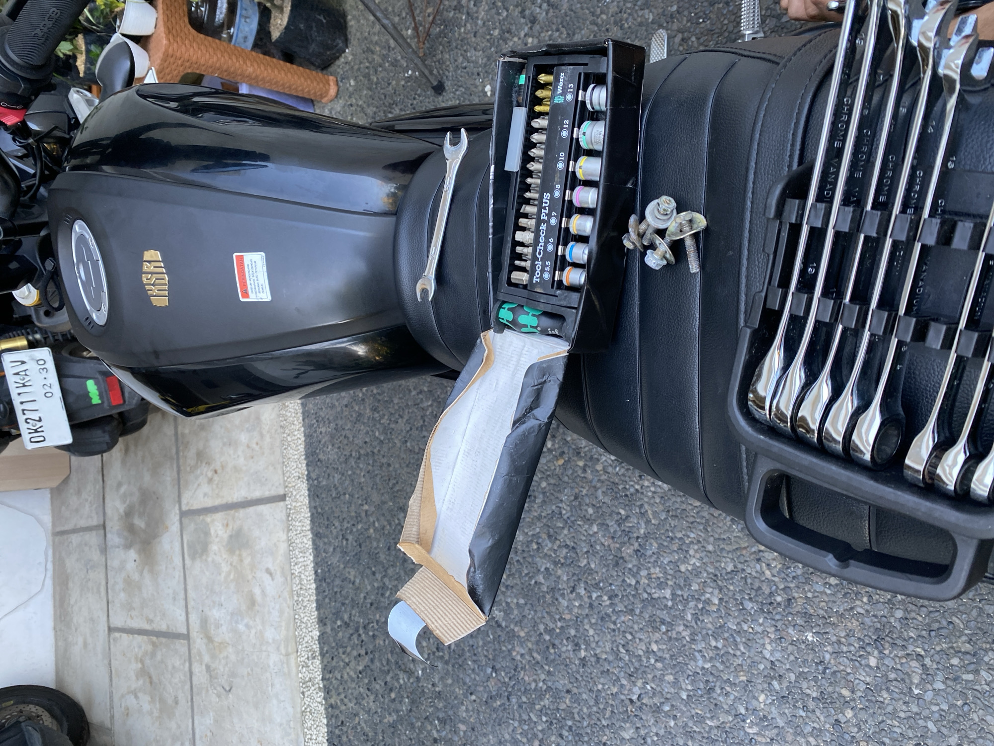 In Kuta Selatan, Indonesia, a black motorcycle is parked on a textured gray asphalt surface, with a tiled pathway visible in the background. Resting on the motorcycle's seat is a black toolbox containing a set of various bits and sockets, labeled "Tool-Check PLUS" and "Wera". Adjacent to the toolbox, a single wrench lies on the seat. A small pile of nuts and bolts is also present on the seat. The motorcycle has a license plate that reads "DK 2711 KAV". The scene appears to be outdoors, with some foliage and potted plants visible in the background.