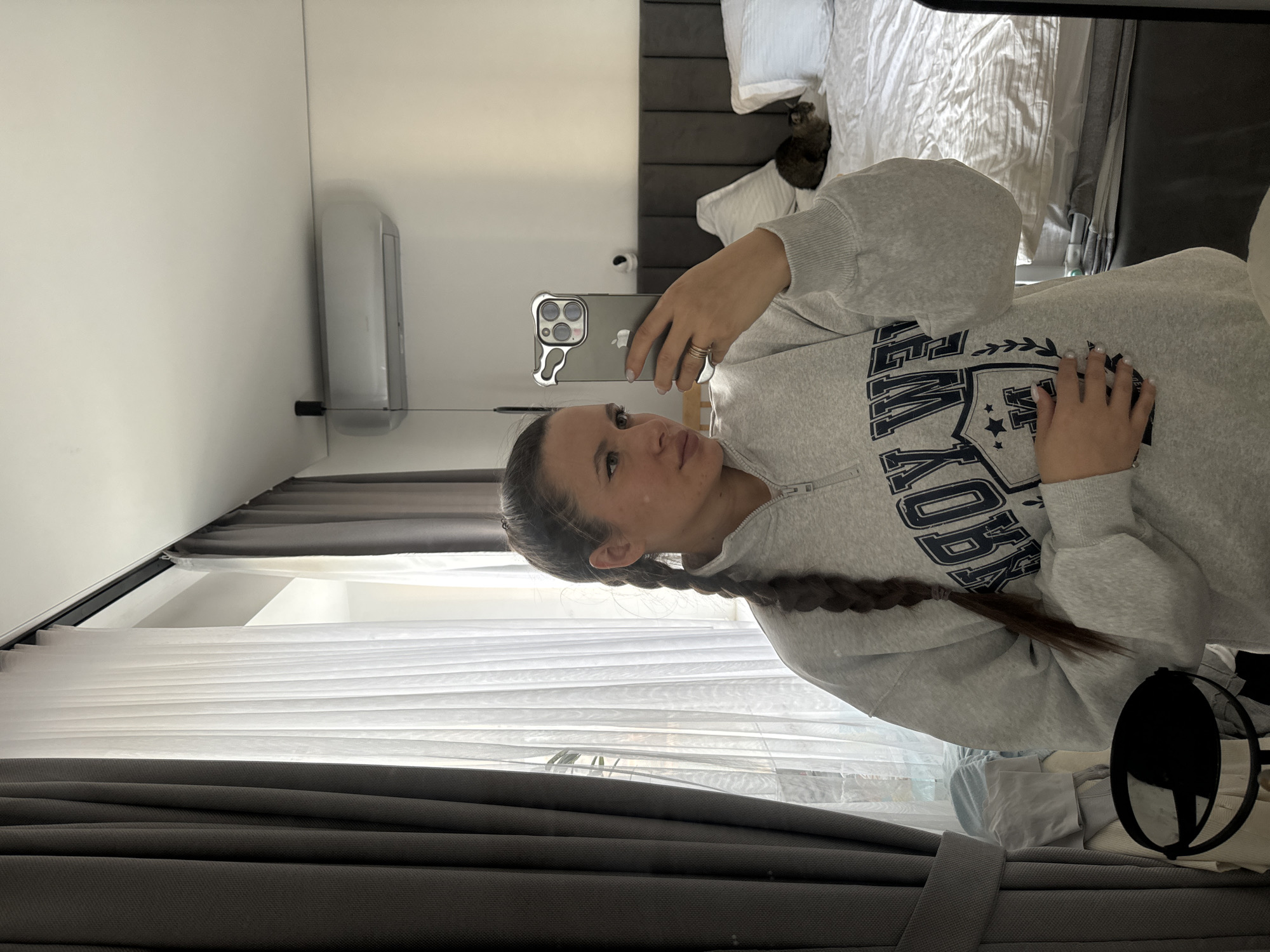 A woman with dark hair styled in a braid is taking a mirror selfie in what appears to be a bedroom. The room includes a bed with white linens and a gray headboard, an air conditioning unit on the wall, and a window with sheer white curtains and gray drapes. A cat is visible on the bed. The specific location within the city of Odintsovo, Russia, cannot be determined from the image.