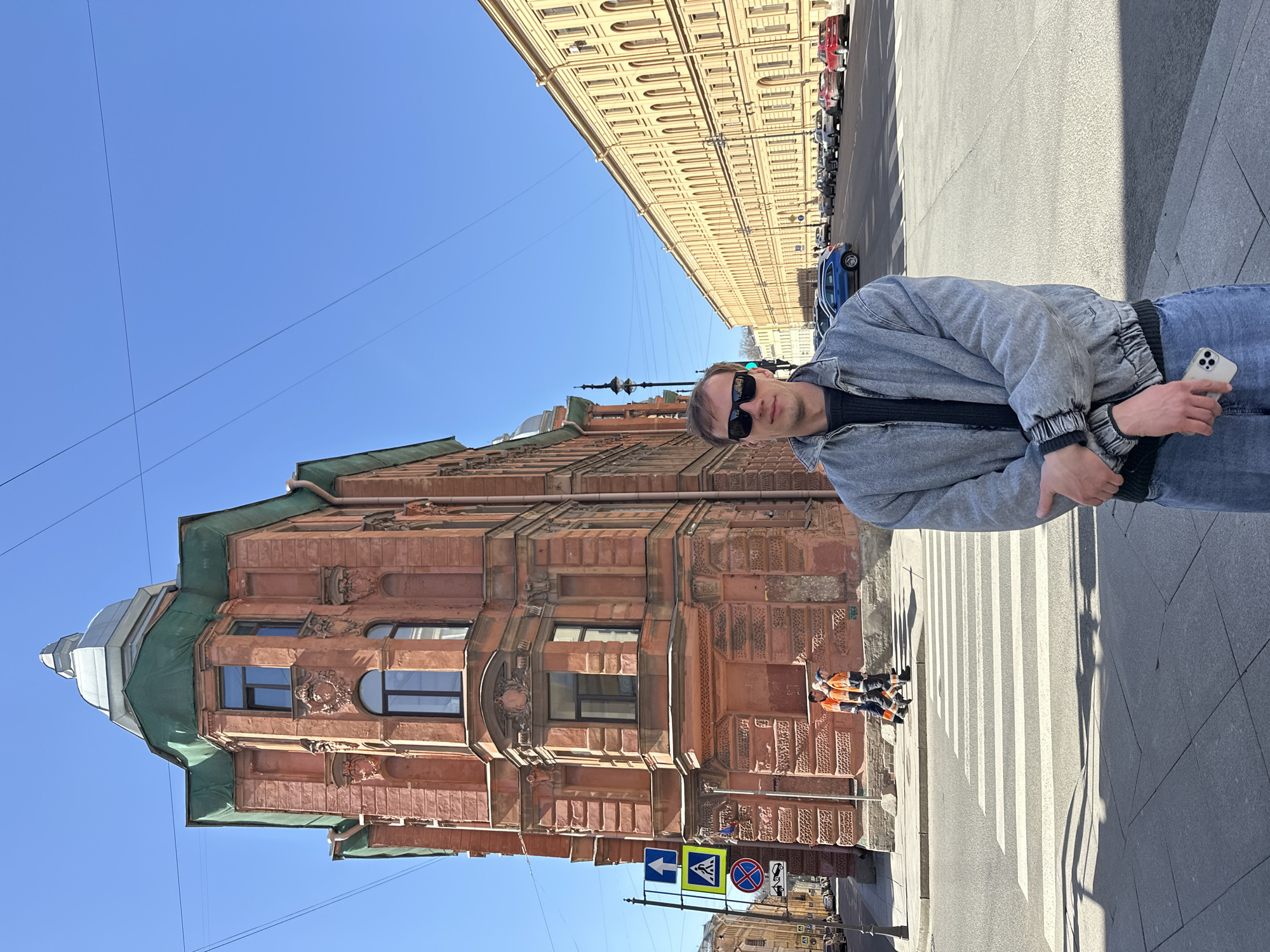 A young man stands on a city sidewalk under a clear blue sky on a bright, sunny day. He is positioned slightly to the right of the frame's center, facing directly towards the viewer. He wears dark sunglasses, a light gray denim jacket over a dark sweater, and blue jeans, holding a white smartphone in his left hand.
Behind him and to his left stands a prominent, ornate reddish-brown building with a distinct green roof and elaborate architectural details, including various window shapes and decorative stonework. Behind him, to his right, is an asphalt street featuring a well-marked pedestrian crosswalk. Several cars, including a red and a blue one, are parked along the far side of the street, which is lined by a long, light-yellow building with many windows.
Near the pedestrian crossing, closer to the reddish-brown building, two individuals in orange and blue work uniforms are visible. Several traffic signs, including a blue arrow, a pedestrian crossing symbol, and a 'no stopping/parking' sign, are posted by the street. The bright daylight casts distinct shadows on the sidewalk to the man's right, indicating midday or early afternoon. The overall scene captures an urban environment with historic architecture, consistent with Saint Petersburg, Russia. No specific text is clearly legible on buildings or signs.