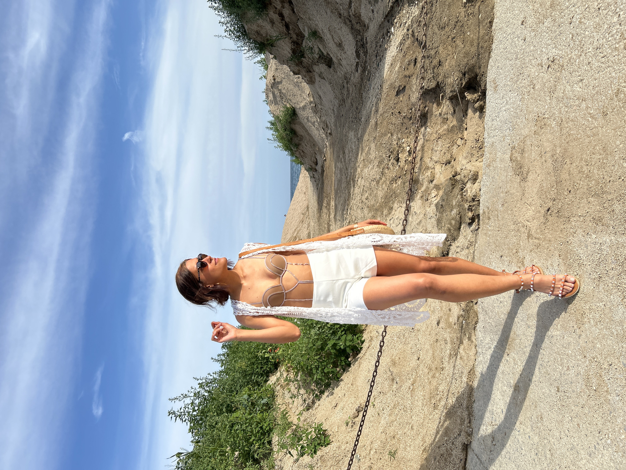 The image depicts a female subject standing on a paved path in an outdoor, sunny coastal environment, identified as Calella, Spain. The woman, with short brown hair and dark sunglasses, faces right in profile, seemingly looking towards the distant horizon. She wears a light brown strappy bralette with white or silver embellishments, white high-waisted shorts, and a long, sleeveless, white crochet duster cardigan. Her feet are adorned with light brown multi-strap flat sandals featuring small white studs. She carries a light brown woven straw bag over her left shoulder, partially visible.
To her immediate left and extending into the midground, a sandy embankment is densely covered with green foliage. To her right, a wider sandy path or gully recedes into the background, flanked by sandy hills with sparse green vegetation. A dark metal chain runs horizontally across the sandy ground in the foreground, bordering the paved path and continuing into the midground. In the far background, a sliver of blue sea is visible on the horizon beneath a light blue sky featuring scattered wispy white cirrus clouds. Long shadows are cast on the paved path to the left of the woman, indicating a sun position to her right.