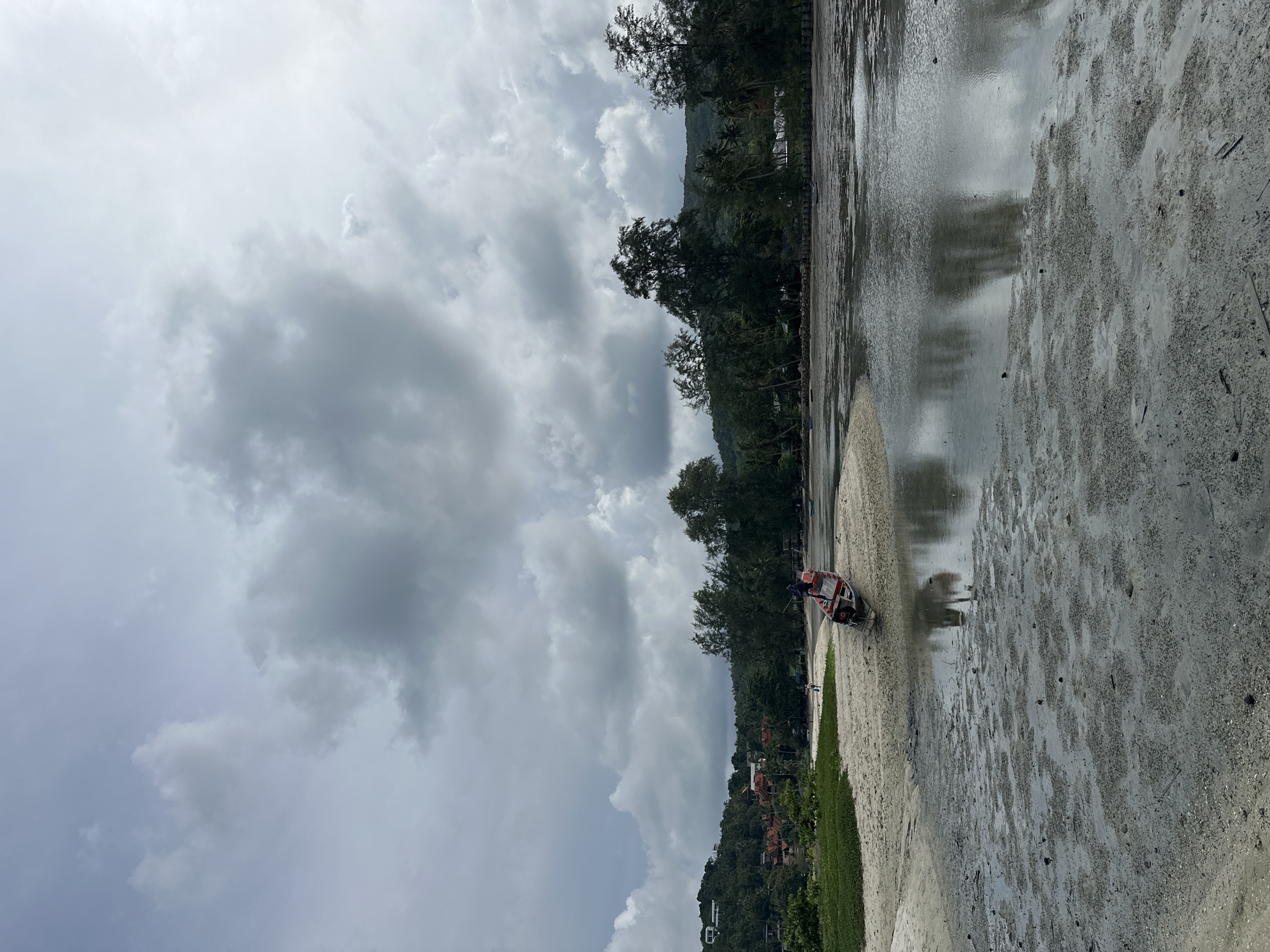 The image depicts a coastal scene on Koh Phangan, Thailand, under a heavily overcast sky with dramatic cloud formations. The foreground is a tidal flat, composed of wet sand and mud with visible ripples and debris, reflecting the muted sky. A single longtail boat, red and white with an outboard motor, rests partially on the sandbank, its reflection visible in the shallow water. In the middle ground, a sandy beach curves to the left, with a patch of lush green vegetation. Further inland, a densely forested hillside rises, dotted with buildings and houses. Trees, including palms and pines, line the coast and the hillside, with a few more small boats visible further down the beach. The overall impression is one of a tranquil, possibly post-rain, coastal environment.