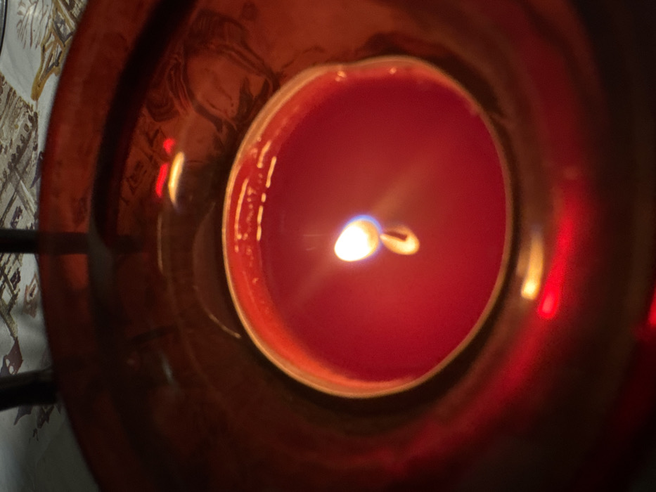Profesor Ishirkovo, Bulgaria: A close-up, overhead view depicts a single red candle burning actively within a dark red, cylindrical glass candle holder. The candle's red wax is partially melted, encasing a visible wick that produces a bright, white-yellow flame with a faint blue base. The flame illuminates the immediate surroundings with a warm, reddish glow. The inner surface of the reflective red glass holder displays several distinct reflections: a faint, distorted profile of a human head or face is visible on the right side, along with two distinct, parallel horizontal red light streaks on the upper left. Subtle reflections of a light-colored patterned textile, consistent with a tablecloth, are also visible. The background, outside the immediate vicinity of the candle holder, is out of focus, showing sections of a patterned textile surface and dark, vertical elements, possibly furniture legs. No direct human presence or interaction is observed beyond the reflection.