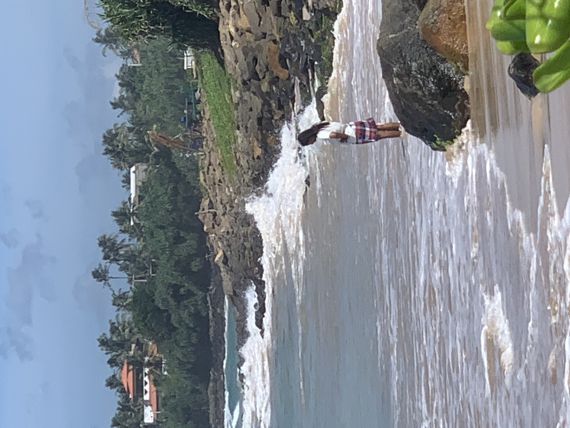 A medium shot shows a woman standing in shallow water at a beach in Kathaluwa, Sri Lanka. She is facing away from the camera, looking out at the ocean. The woman is wearing a white t-shirt and a red and white plaid skirt. She has dark hair. Waves are breaking on the shore and around large rocks in the water. Behind the beach, there is a rocky embankment covered in green vegetation, with palm trees and buildings visible in the distance. The sky is cloudy. In the foreground, out of focus, are bright green leaves of a plant.