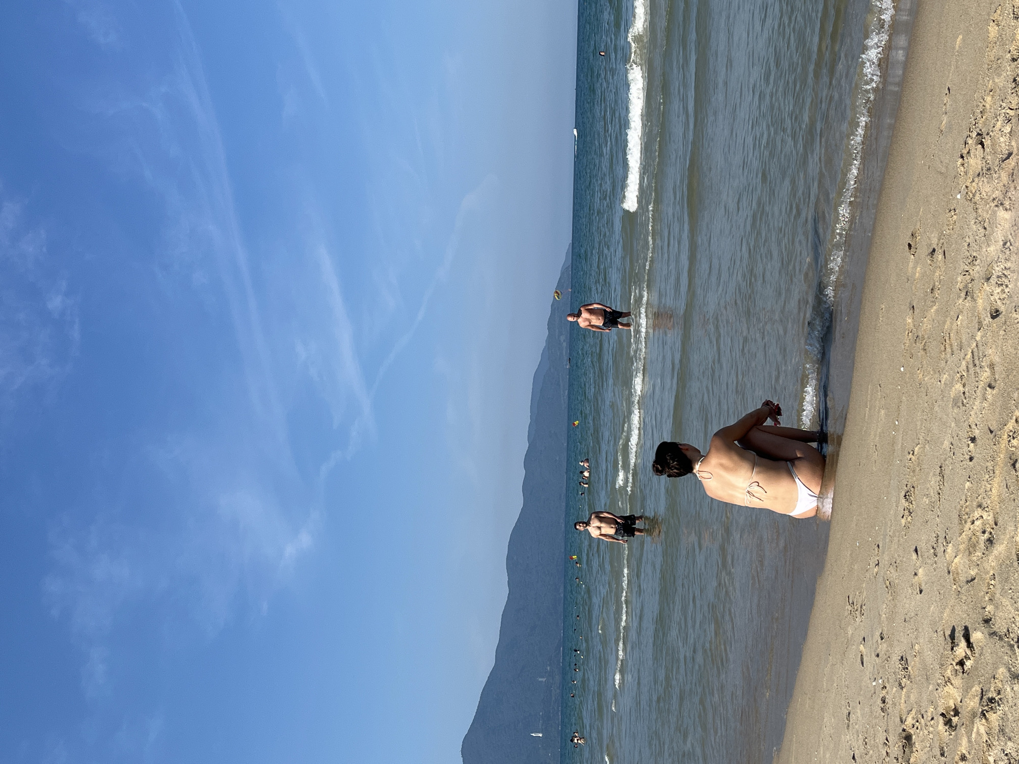 The image shows a beach scene in Đà Nẵng, Vietnam. The sky is blue with thin white clouds. In the ocean, there are several people wading in the water. A woman in a white bikini is sitting at the edge of the beach with her back to the camera. The beach is sandy. A mountain range is visible in the background.