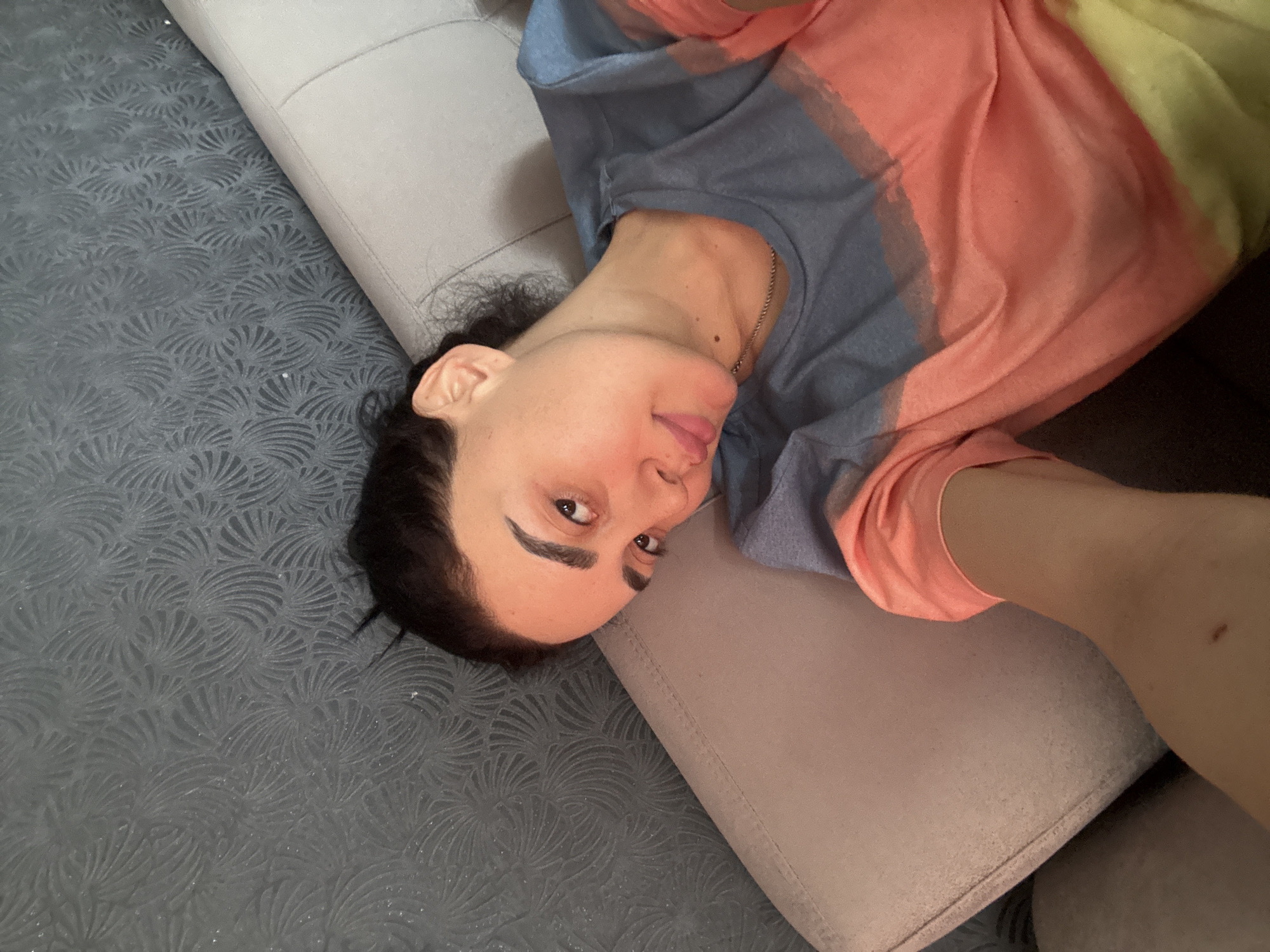 A person is lying on a couch. The person has dark hair and is wearing a multicolored shirt. The couch is upholstered in a light gray fabric. The background is a dark gray patterned carpet. The location is not discernable from this image.