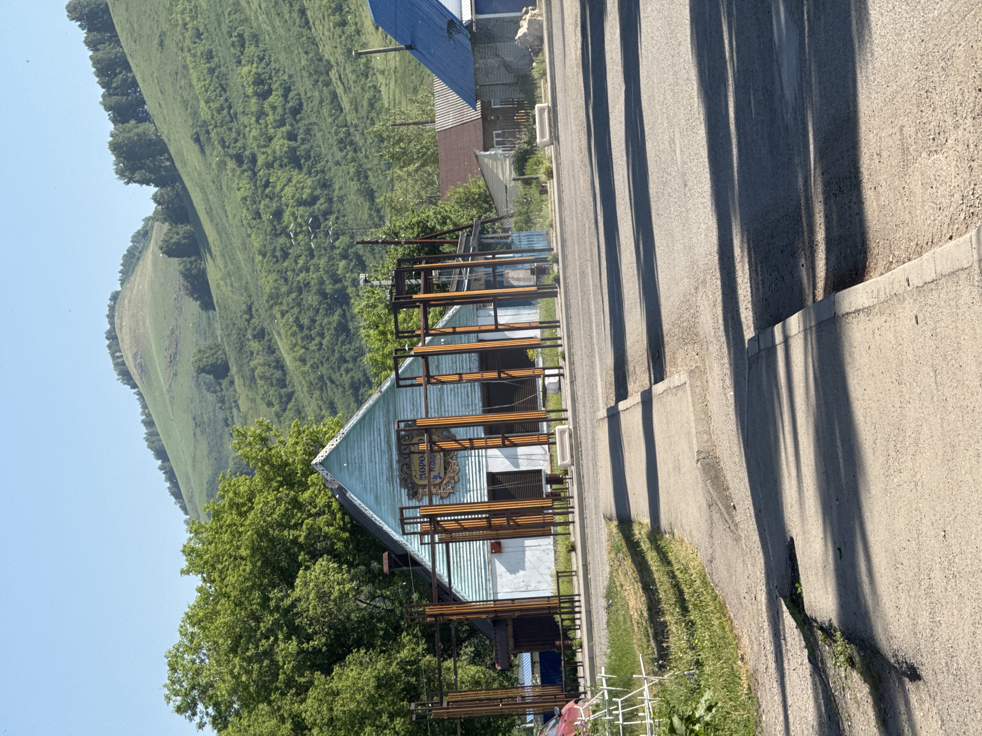The image shows a street scene in Soloneshnoye, Russia, on a sunny day. In the foreground, a paved road and sidewalk are visible, with shadows cast by unseen objects. A large pothole is present on the road's edge. Behind the sidewalk, a building with light blue siding and a white lower half stands, featuring a prominent sign with Cyrillic text that appears to read "МОРОЗКО" (Morozko), likely indicating a shop or cafe. This building is framed by a series of vertical metal structures with orange accents. To the right of this building, two other houses are partially visible: one with a reddish-brown corrugated metal roof and a yellow-green facade, and another with a blue metal roof. Behind the buildings, a lush green hillside rises, covered with trees and vegetation. The sky is clear and blue. A red car is parked on the left side of the image, near a white wire structure.