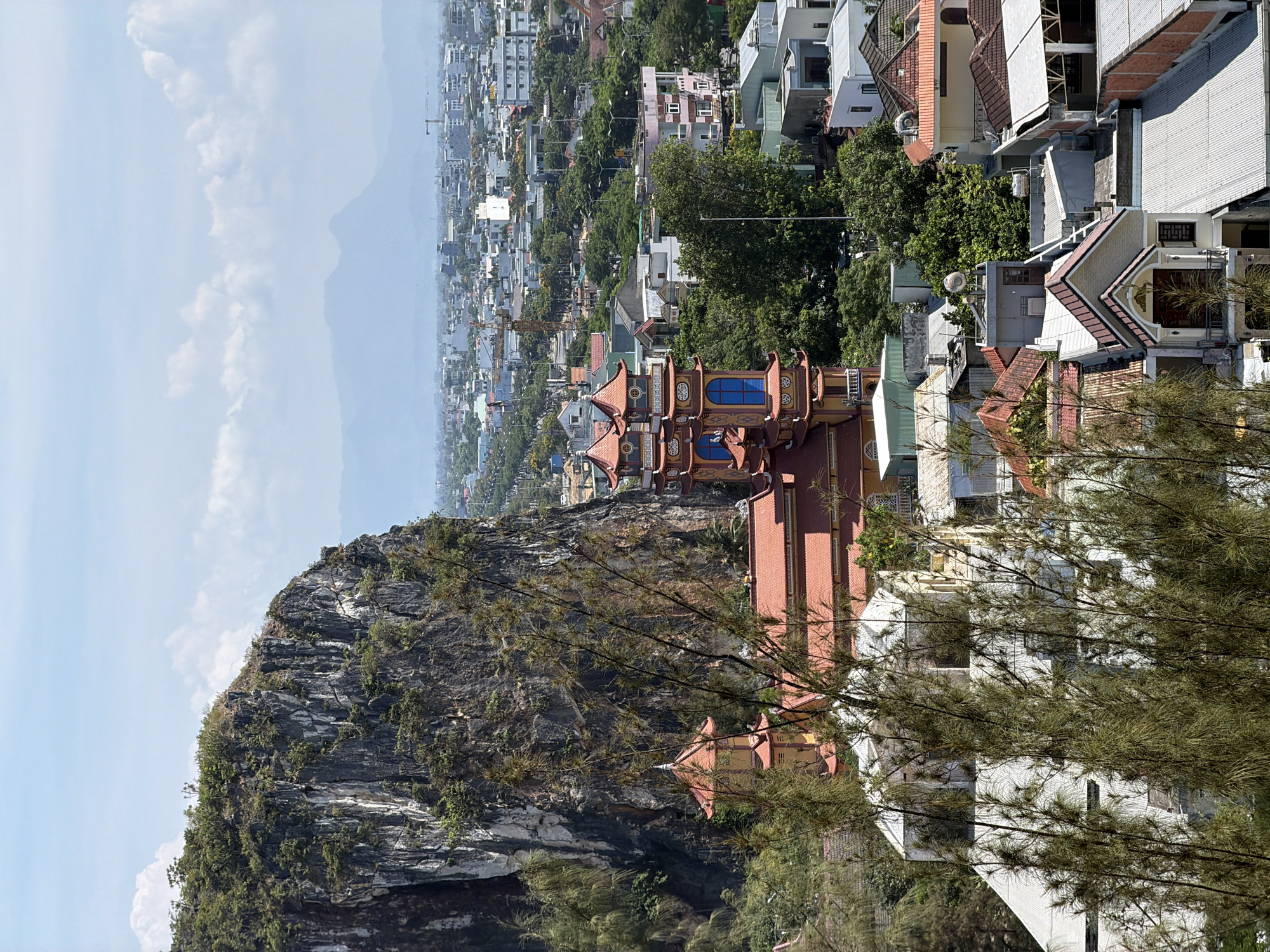 A horizontally oriented, eye-level shot captures the cityscape of Đà Nẵng, Vietnam. A tan and red pagoda stands to the right, with its architectural lines and blue windows contrasting against the greenery. Behind it, urban dwellings form the backdrop, their rooftops and structures interwoven with trees. To the left, a rock formation covered in vegetation rises, adding a natural element to the scene. The sky, filled with soft clouds, meets the distant urban skyline on the horizon. The foreground features thin, green foliage, partly obscuring the view.