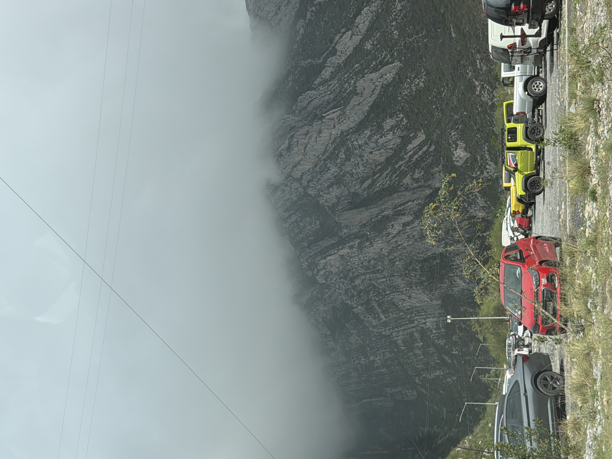 The image captures a mountainous landscape partially shrouded in fog, with a line of vehicles parked on the right side of the frame. A large, rugged mountain or cliff face, textured with rock and sparse vegetation, dominates the background, with a thick layer of fog or cloud obscuring its upper and central portions.
In the foreground, a row of cars is parked facing to the left, perpendicular to the main image orientation (which is rotated). Prominently visible are a red car, followed by a bright yellow-green off-road vehicle (likely a Jeep), a white pick-up truck or SUV, a dark grey SUV, and a black SUV at the end of the line. These vehicles appear to be parked on a paved or gravel roadside. To the right of the parked cars, there's a strip of grassy and rocky ground with small bushes. A few light poles or street lamps are visible behind the parked cars. Overhead, multiple thin lines, possibly power lines or cables, stretch across the pale, overcast sky on the left side of the image.
The weather appears foggy or cloudy, especially impacting the visibility of the mountain. The lighting suggests it is daytime, but overcast. There is no discernible human activity or visible text in the image. The scene is set in Santa Catarina, Mexico, featuring a natural and developed roadside environment.