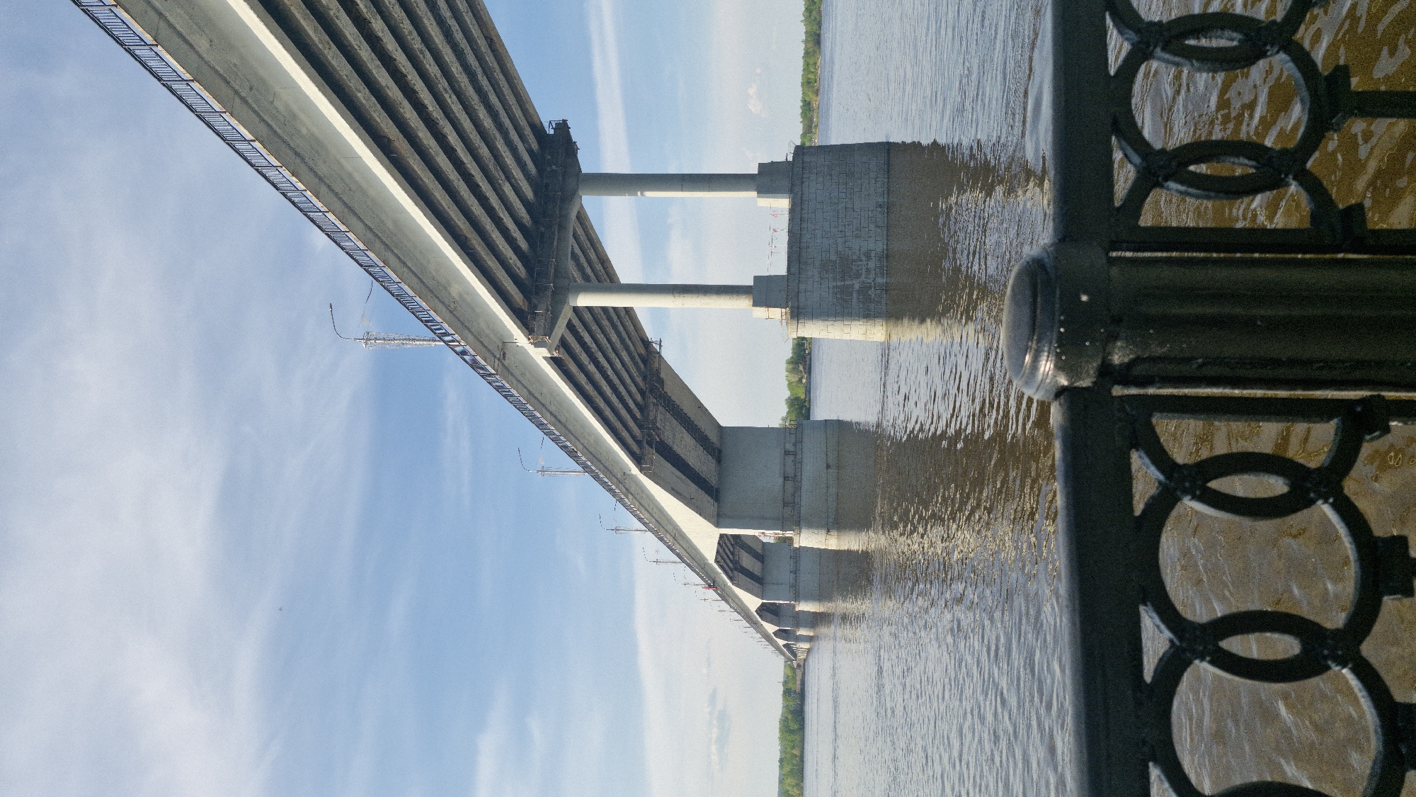 A low-angle shot of a large concrete bridge spans a wide, muddy river under a bright blue sky with some wispy clouds. The bridge's support pillars are visible in the river. The riverbank with green vegetation is in the distance. A decorative metal railing runs along the edge of the frame, partially obscuring the river. This is the Kommunalny Bridge across the Kama River in Perm, Russia.
