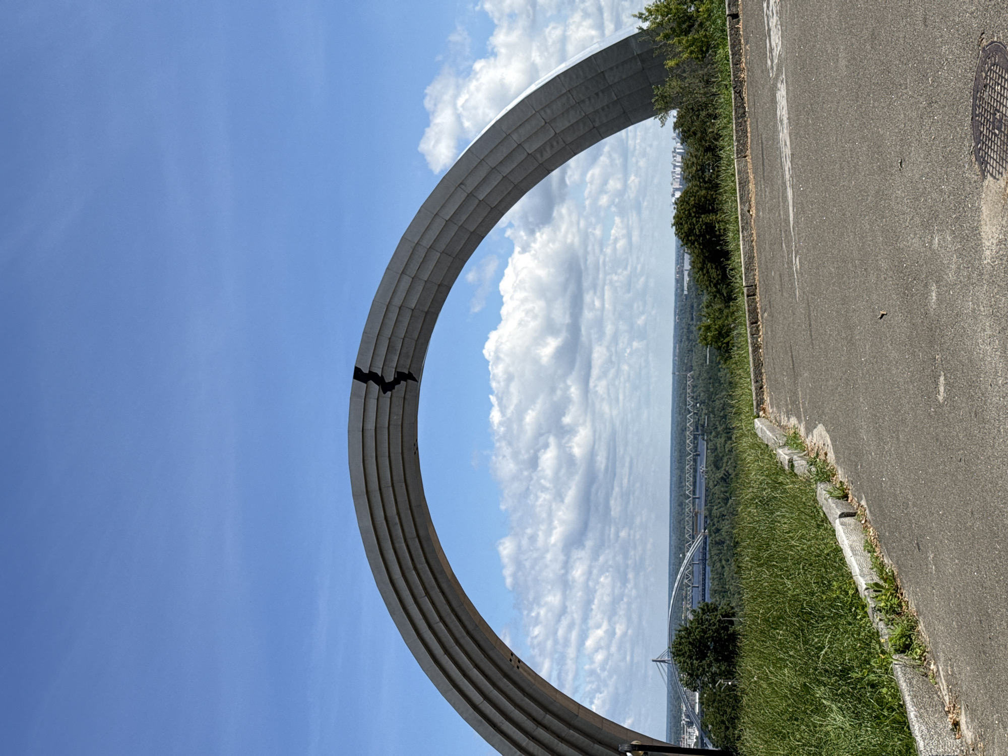 This is a photograph of the Arch of Freedom of the Ukrainian People, formerly known as the Friendship of Peoples Arch, in Kyiv, Ukraine. The monument is a large, steel archway that spans across a portion of the sky. The arch is made of several curved sections, with a dark, jagged line running through the center of the upper portion. In the foreground, there is a paved road with a manhole cover visible. To the left of the road, there is a grassy slope with some rocks and vegetation. In the background, there is a panoramic view of the city of Kyiv, including the Dnipro River and several bridges. The sky is blue with scattered white clouds. There are no people visible in the image.