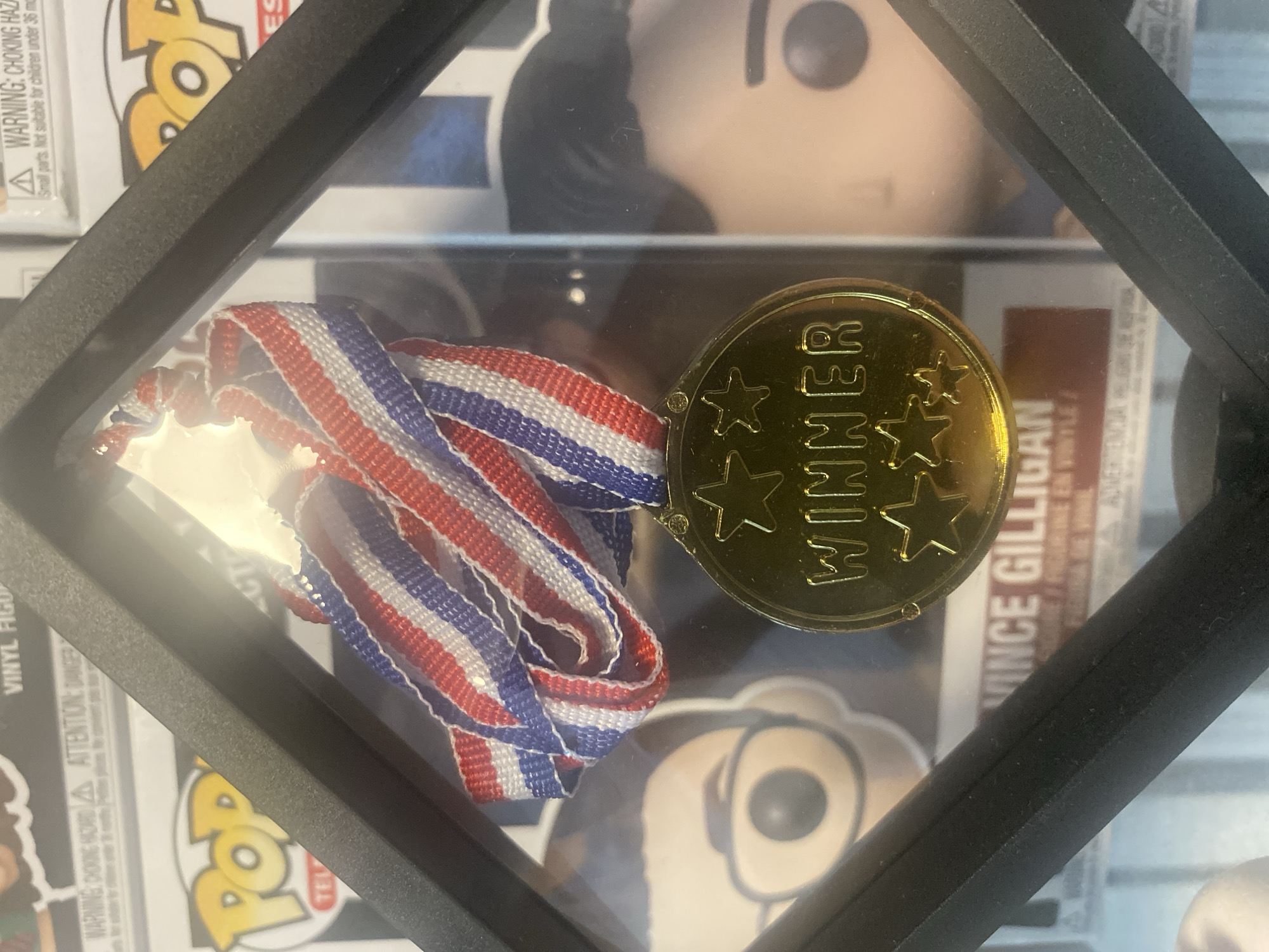 The image shows a framed gold medal with a red, white, and blue ribbon. The medal is inscribed with the word "WINNER" and three stars. It is displayed in front of several Funko Pop! figures, including one labeled "VINCE GILLIGAN." The background suggests an indoor environment, possibly a collector's display. The image does not provide any information about the location in the city of Kamianske, Ukraine.
