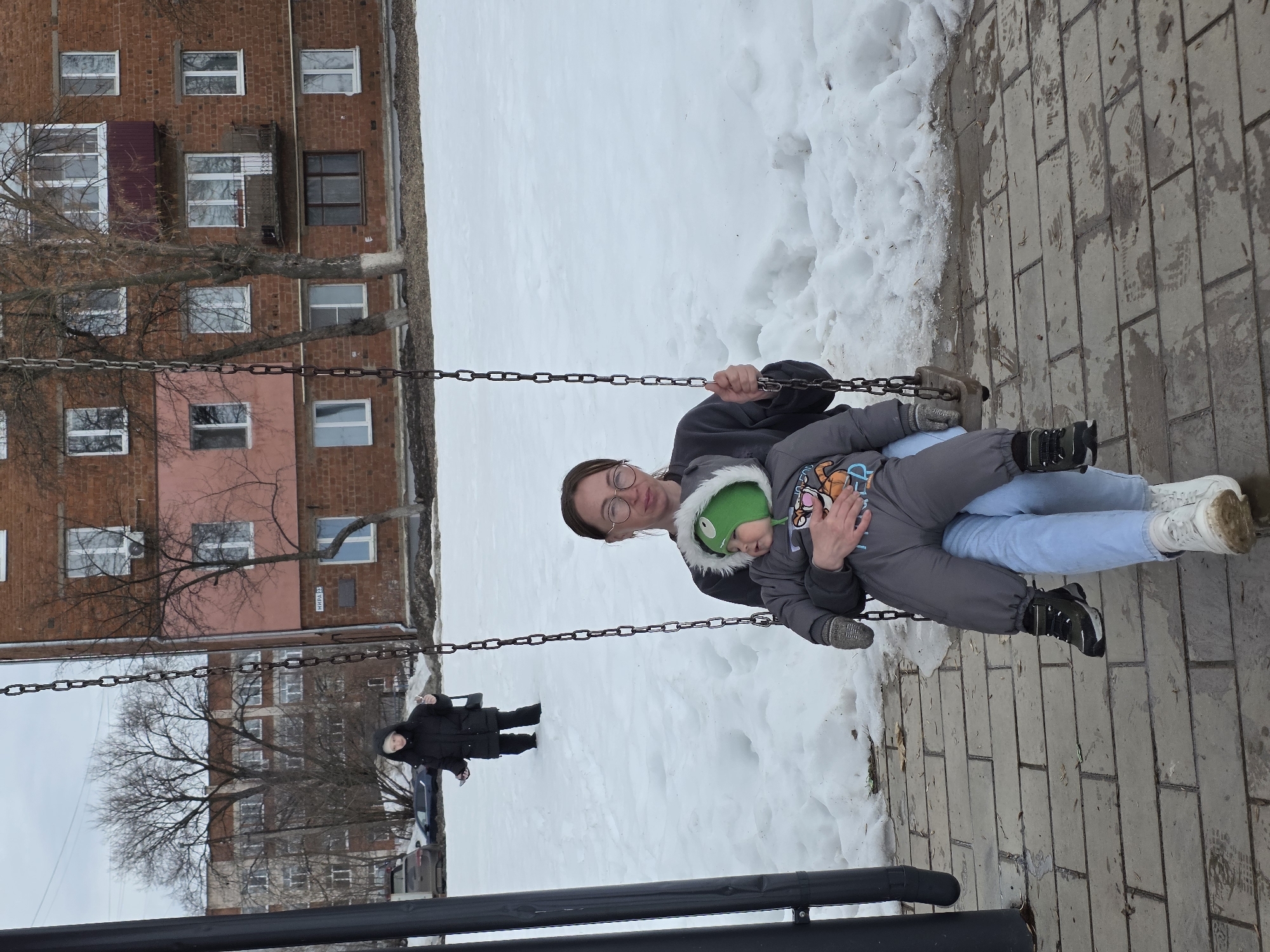 A woman wearing glasses and a dark gray sweatshirt is on a swing with a young child wearing a gray snowsuit and green hat. The child is lying in the woman's lap. The background consists of snow-covered ground, bare trees, and brick apartment buildings. A person in a black coat stands in the background. The ground in the foreground is paved with gray rectangular blocks. Based on visual cues, the image is likely taken in Votkinsk, Russia.