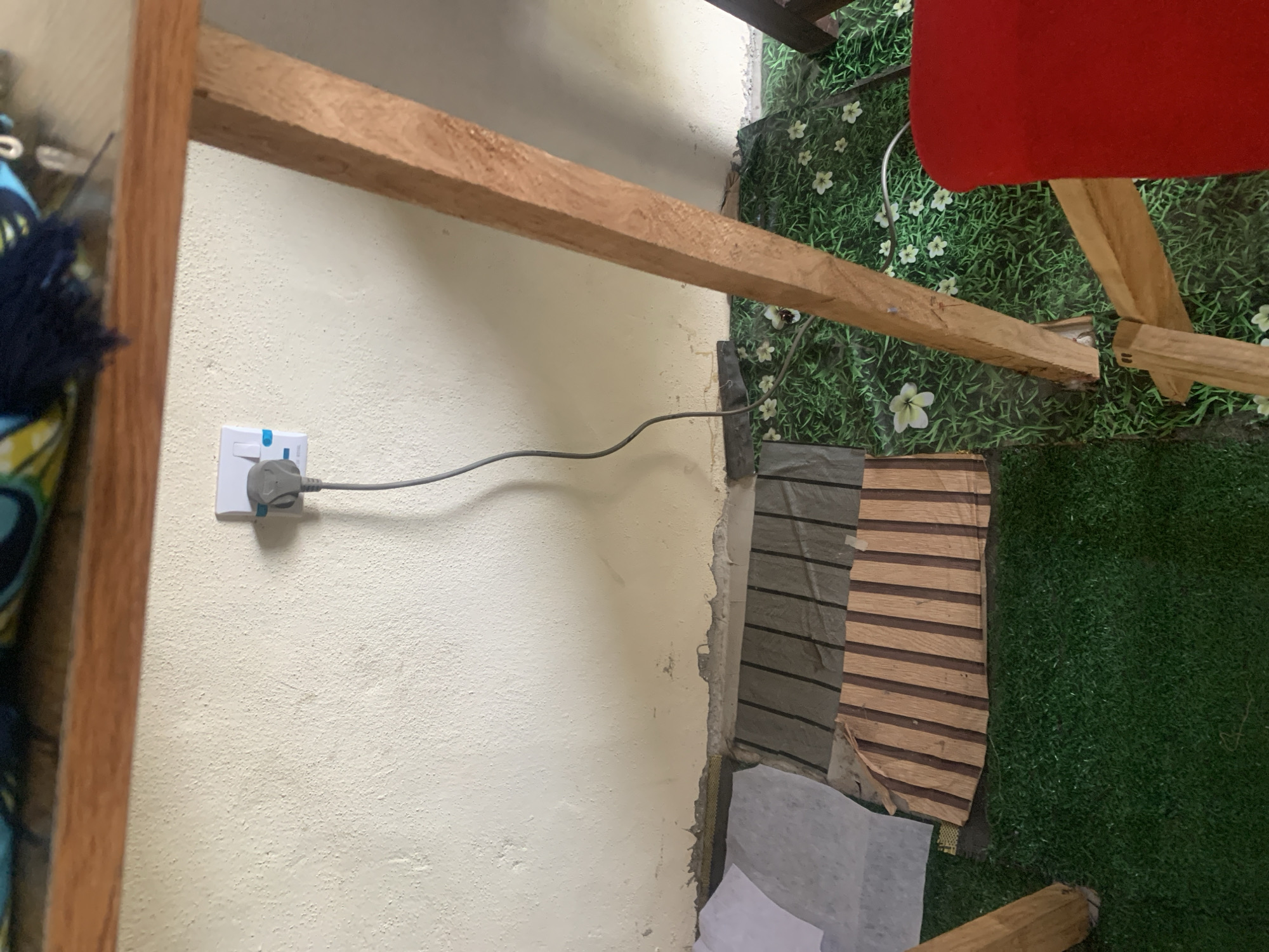 The image shows a corner of a room with a light-colored textured wall and a wooden structure. An electrical outlet with a gray plug is visible on the wall. Below the wall, there's a section of what appears to be artificial grass with a floral pattern, and a wooden mat with a striped design. Part of a red object, possibly a chair, is in the lower right corner. The overall environment suggests an indoor setting, possibly a home or a workshop. The location specified, Jauro Yinu, Nigeria, is not visually discernible from the image itself.