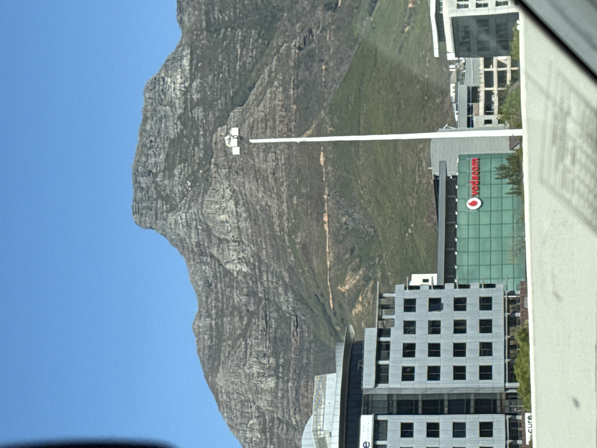 The image shows a mountain in Cape Town, South Africa, with buildings in front of it, the Vodafone building visible. A streetlight is in the foreground. The sky is blue.