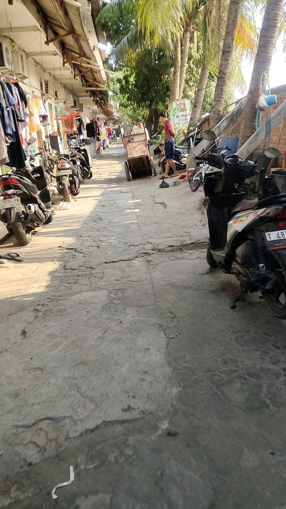 The image depicts a street scene in Mekarmukti, Indonesia, characterized by its weathered concrete ground and buildings lining one side, likely a marketplace or commercial area. On the left, clothes are hung outside a building, suggesting a retail establishment. Several motorcycles are parked along the side of the street.
In the center of the frame, a man wearing a red shirt and dark shorts is pushing a wooden cart. Another person, partially visible, is near the cart. Further down the street, more people and motorcycles are present. Palm trees are visible in the background, indicating a tropical or subtropical environment. The lighting suggests it is daytime, with shadows cast across the ground. The overall impression is one of a busy, everyday street in a developing urban area.