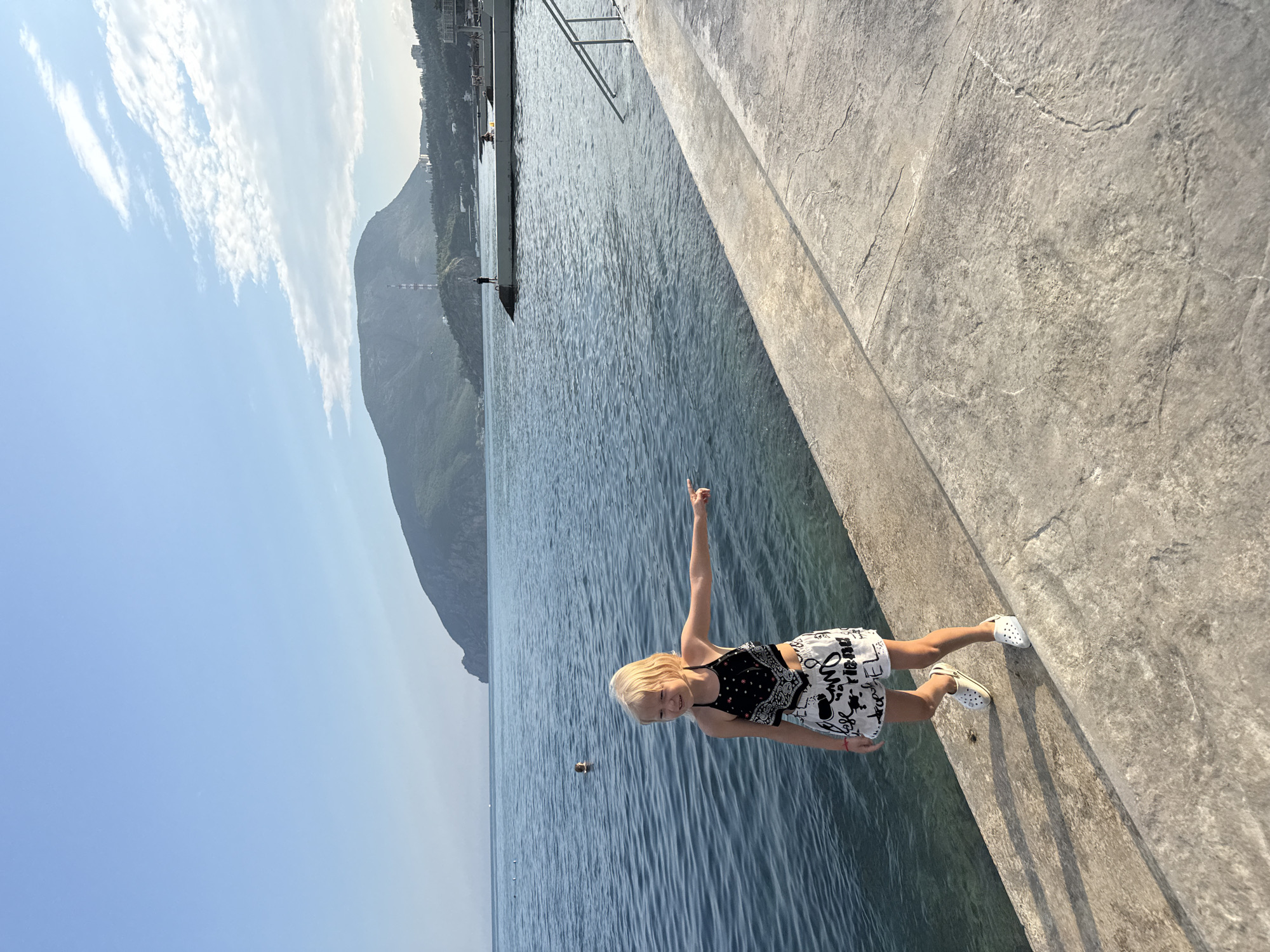 A young blonde girl stands on a concrete embankment overlooking a body of water. She is wearing a black and white patterned halter top and black and white patterned shorts, along with white clogs. Her right arm is extended, pointing towards the water. In the background, a large, mountainous landmass rises from the sea, with buildings and a tall tower visible on its slopes. The water is calm with ripples, and a few small buoys are visible in the distance. The sky is clear with some scattered clouds. The scene appears to be the coastal area of Utios, Ukraine.