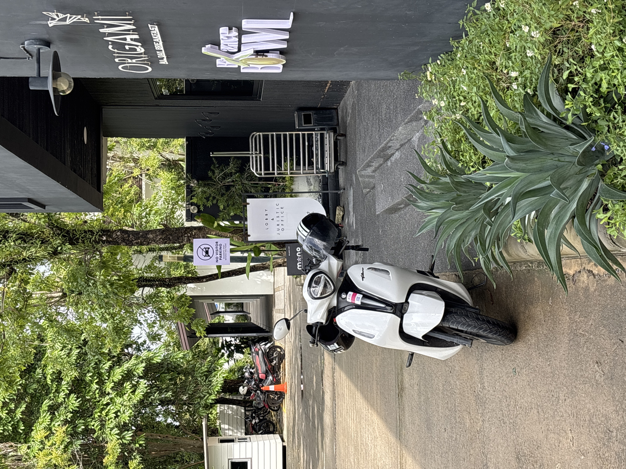 The image depicts a street scene in Choeng Thale, Thailand. A white electric scooter is parked on the right side of the image. A black helmet rests on the scooter. In the background, several other motorcycles are parked. A sign indicating "No Visitor Parking Beyond This Point" is visible. Another sign displays "Lobby & Juristic Office." The building features signage for "Origami All Day Breakfast" and "Cake Bowl." Lush greenery grows along the edge of the building and surrounds the parking area.
