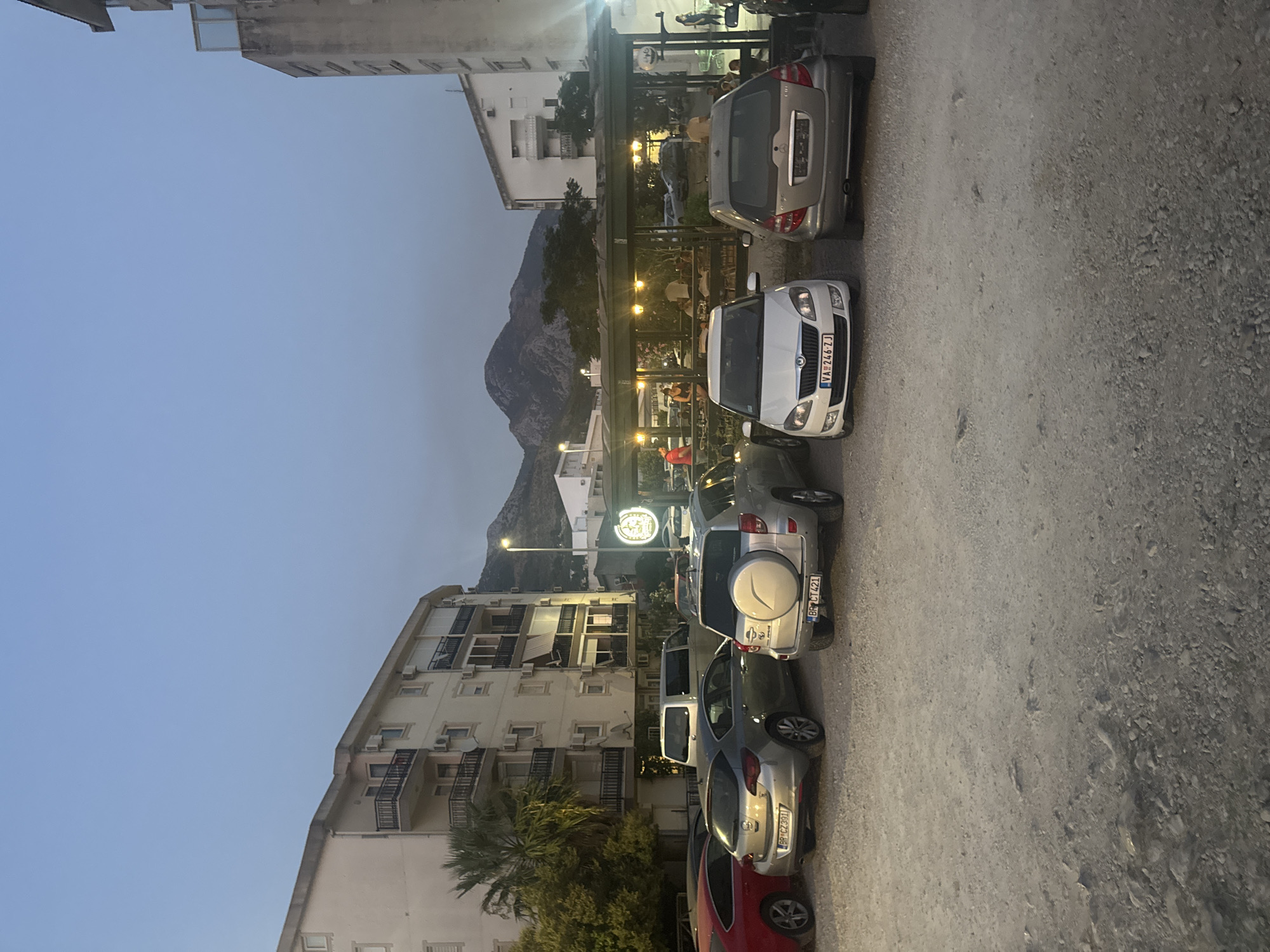 The image depicts an outdoor scene in Sutomore, Montenegro, at dusk. In the foreground, a gravel parking lot contains several parked cars, including a silver SUV with a spare tire mounted on the back, a white Skoda Fabia, and a silver hatchback. Behind the cars, a covered outdoor seating area of a restaurant is visible, illuminated by string lights and lamps. People are seated at tables within this area, some appearing to be dining or socializing. Beyond the restaurant, buildings and a palm tree are visible, with mountains forming the backdrop against a clear, pale blue sky. A street lamp is also present. The overall atmosphere suggests a casual evening setting.
